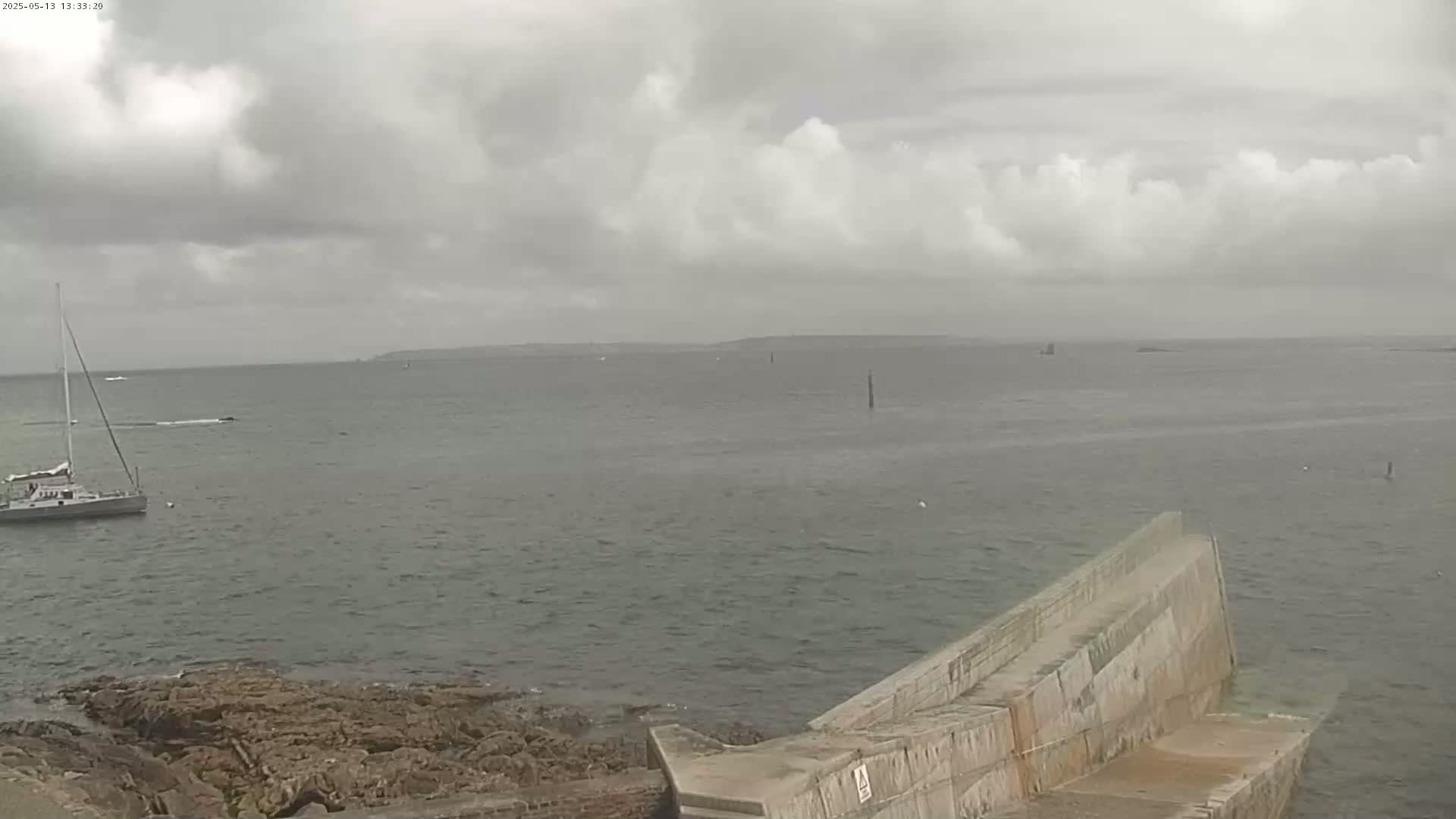 A sailboat is anchored in a calm sea under a cloudy sky near a rocky shore and a concrete breakwater.