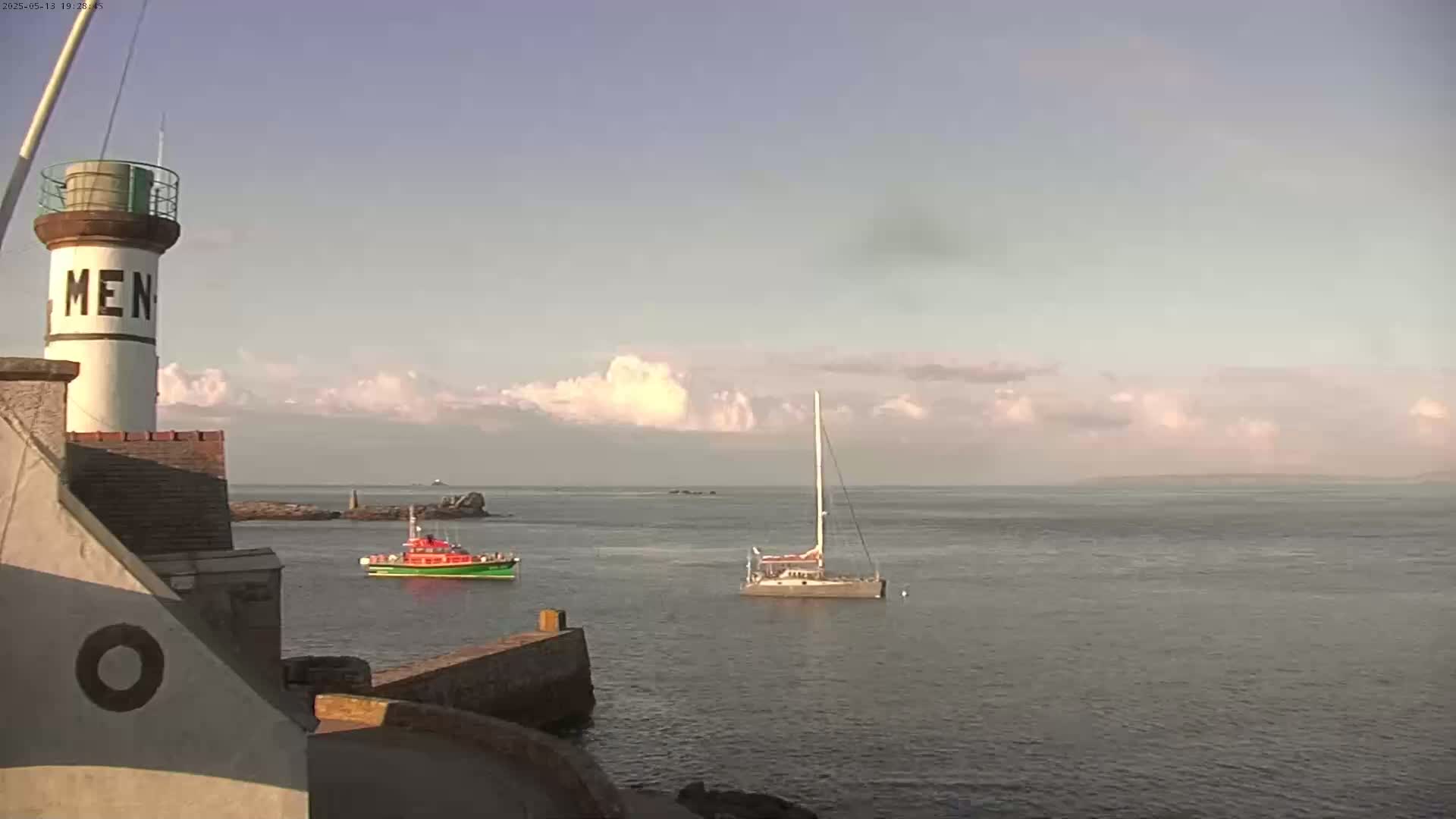 A lighthouse stands near a harbor with a sailboat and a small rescue boat on a calm sea under a partly cloudy sky.