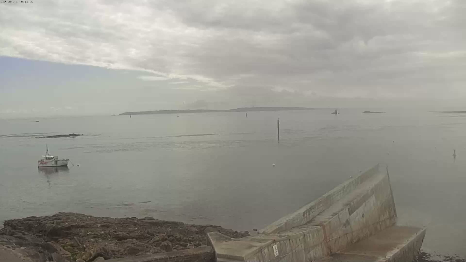 A small boat sits in calm, hazy water near a rocky shoreline and a partially submerged concrete structure, with a distant island visible under an overcast sky.