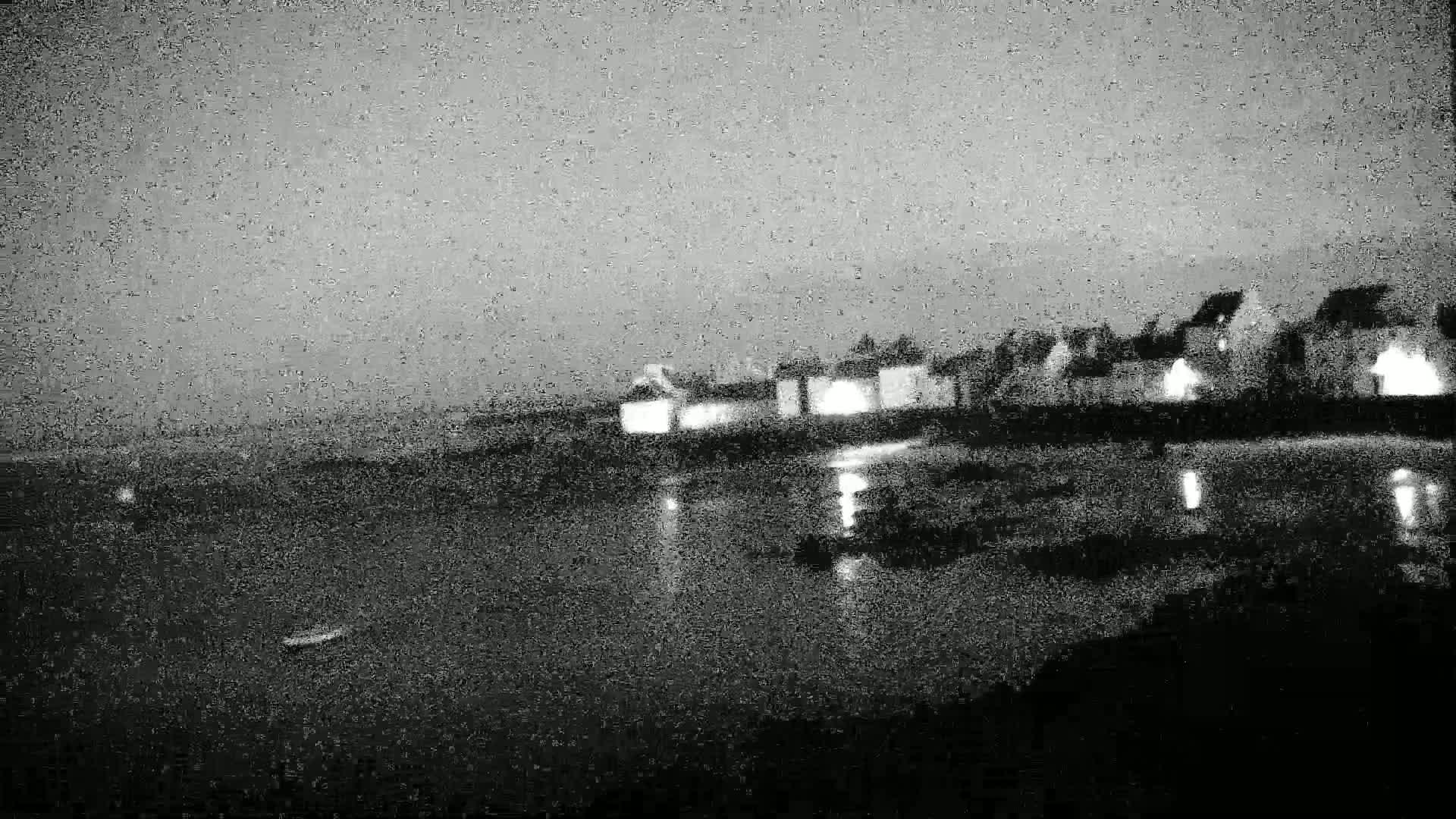 A nighttime, overcast view of a waterfront village with dimly lit buildings reflected in calm water.