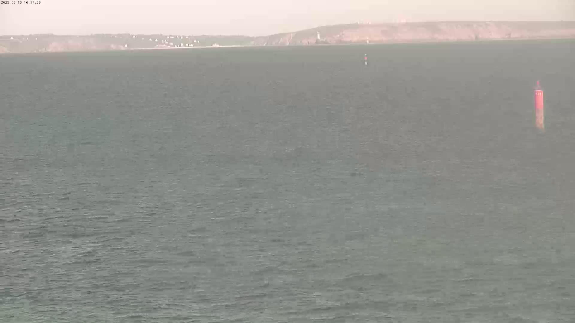 A calm, gray sea stretches to a distant hazy shoreline under an overcast sky, with a single navigational buoy visible in the water.