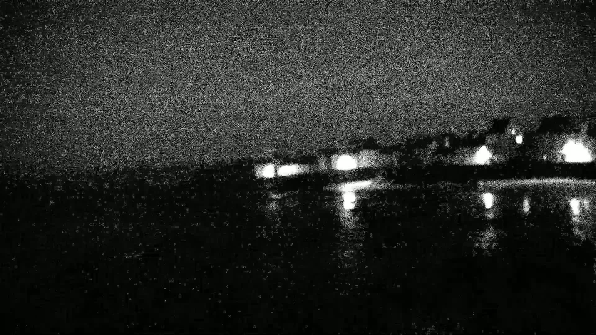 Lights from buildings are reflected in dark water under a grainy, nighttime sky.