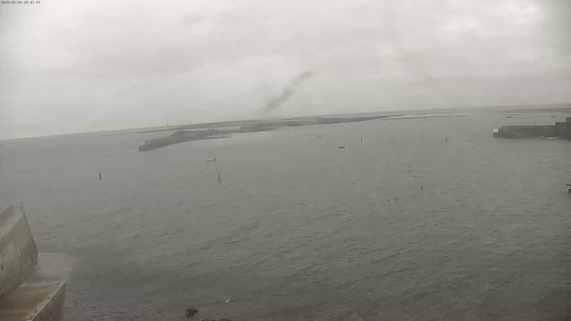 A wide, overcast view of a harbor with several small boats and a plume of dust or smoke rising in the distance.