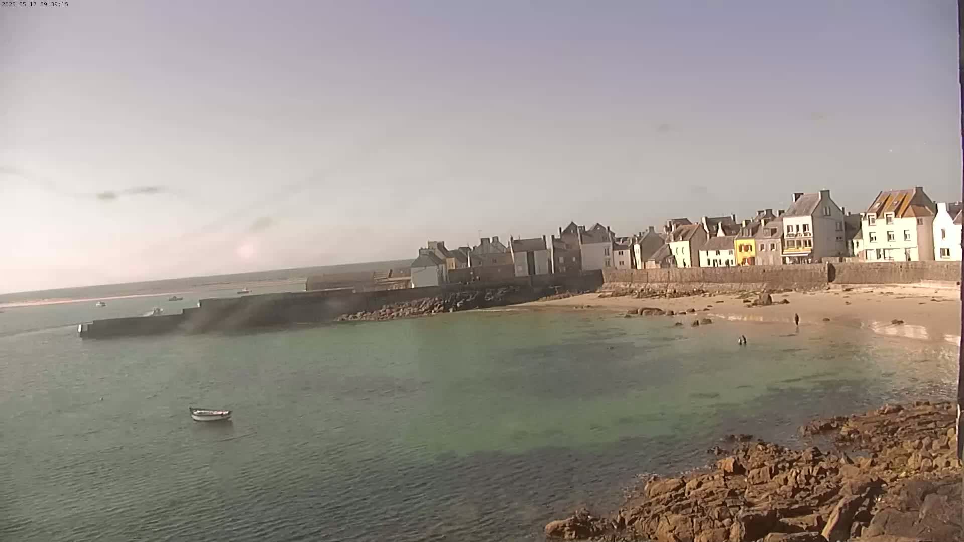 A small, sandy beach with calm, green water sits in front of a row of buildings along a stone wall under a hazy, sunny sky.