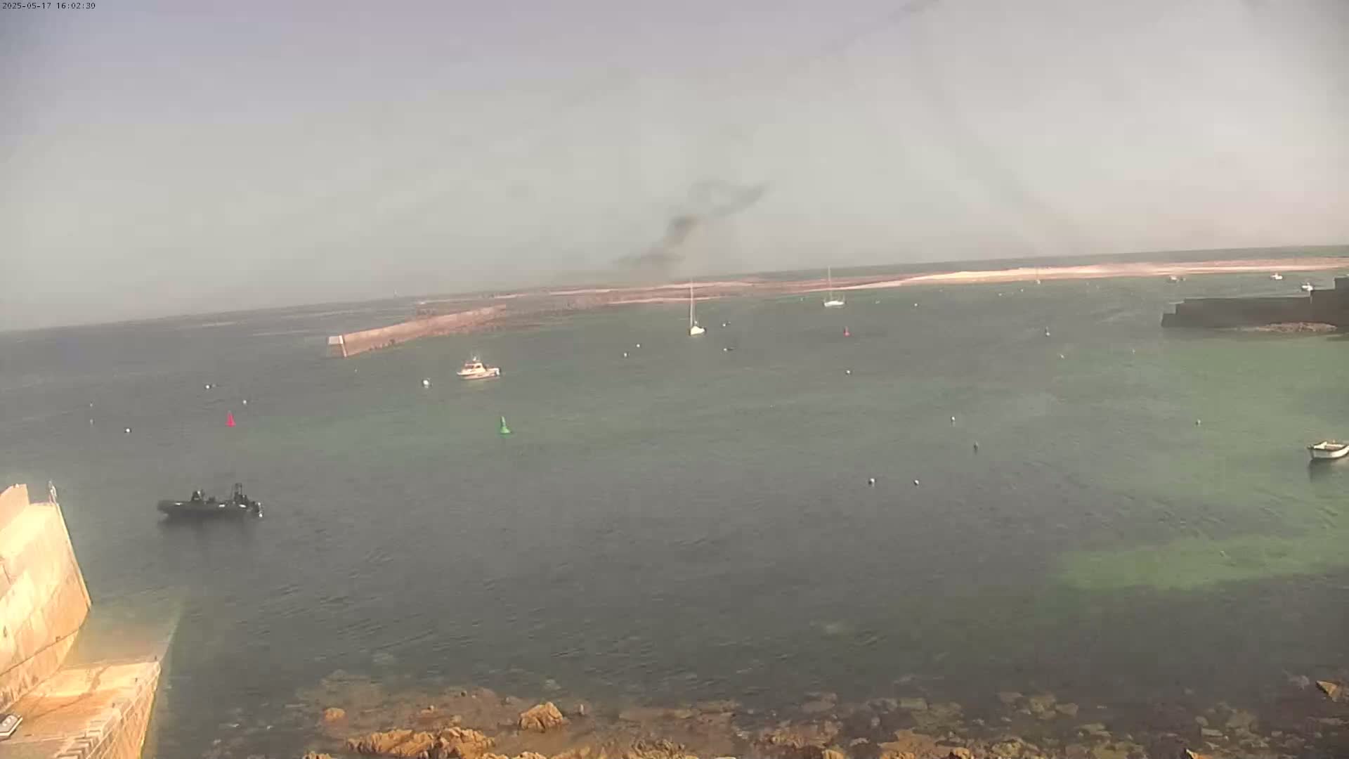 A hazy, sunny day shows a calm harbor with several boats and a small, dark-colored rigid-hulled inflatable boat near a stone wall.