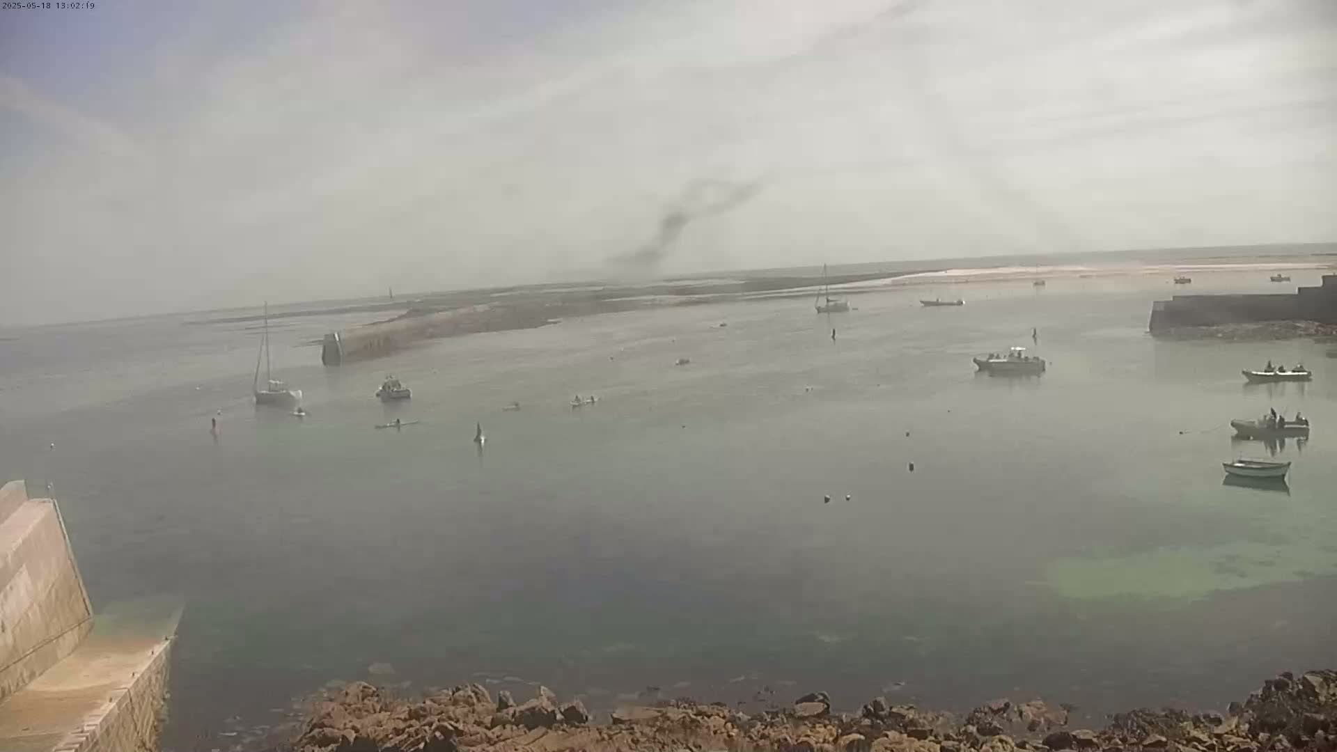 A hazy, calm bay with several small boats and sailboats is seen from a rocky overlook.