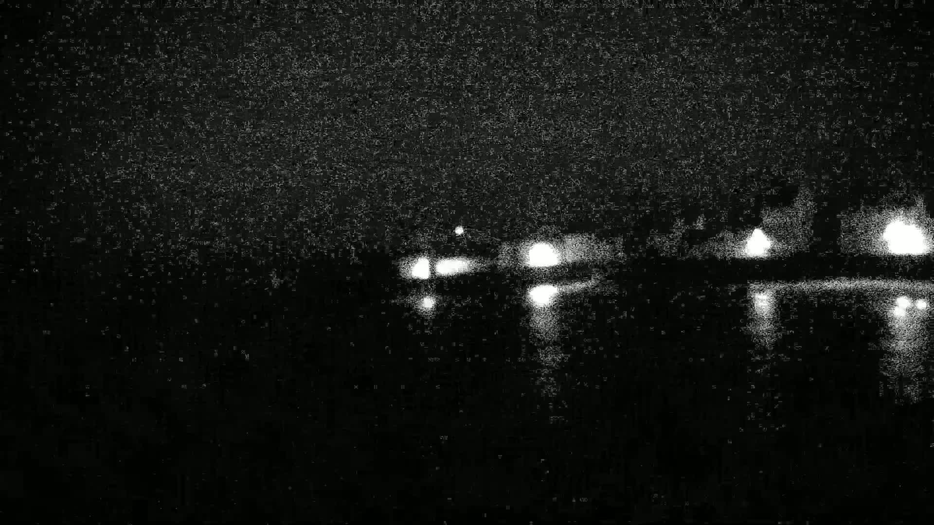 A blurry nighttime image shows the lights of vehicles on a road reflecting on a dark body of water under a snow-filled sky.