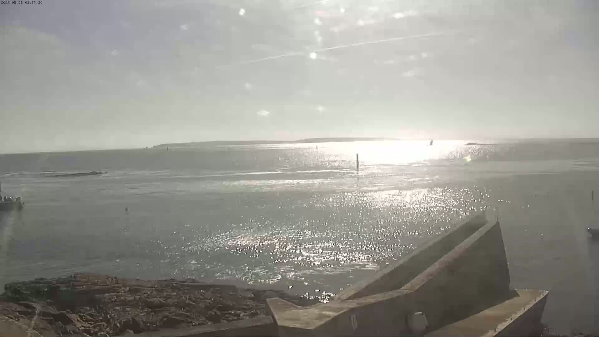 A bright, sunny day overlooks a calm body of water with a distant shoreline, viewed from a rocky coast with a concrete structure in the foreground.