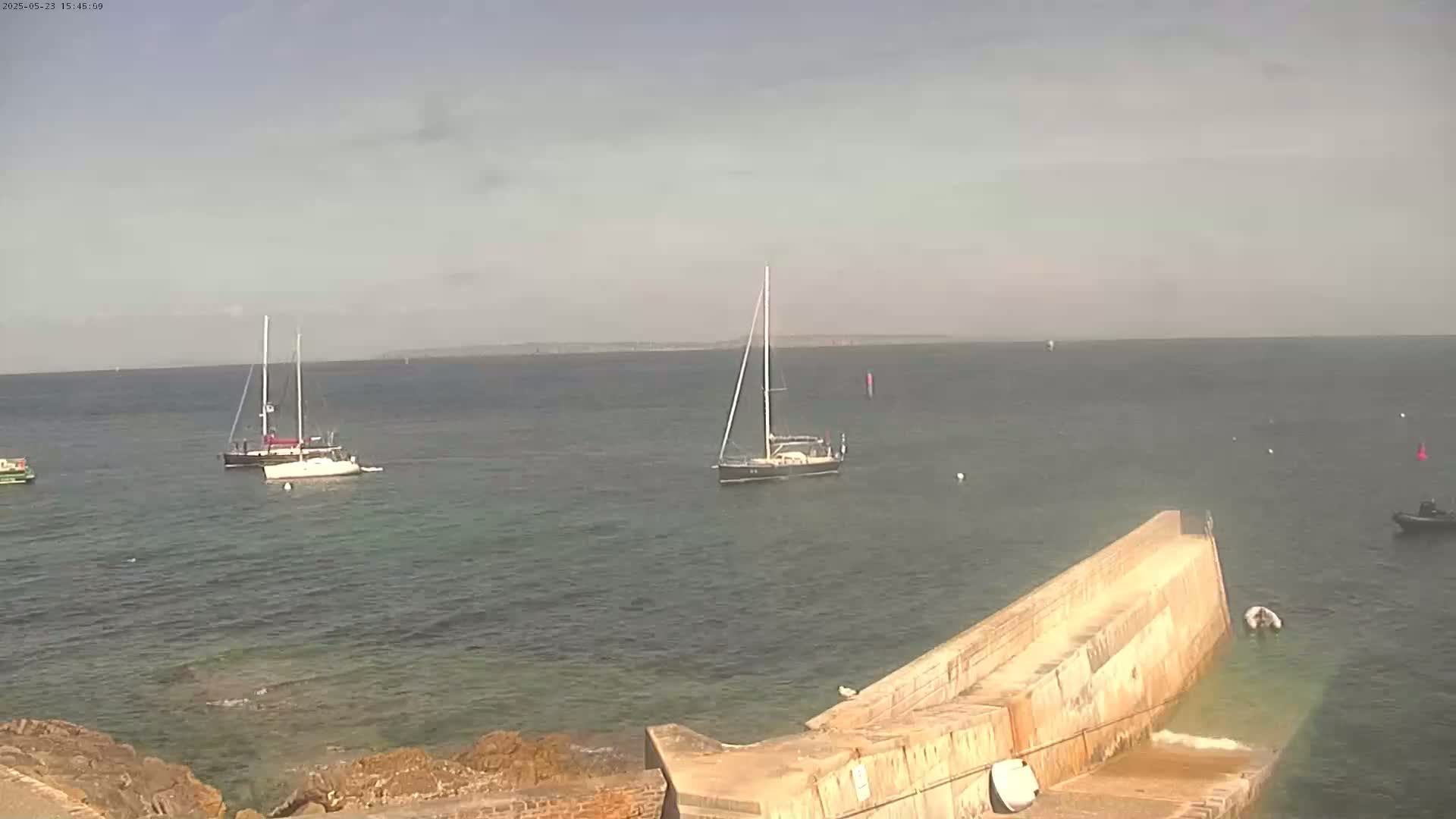 Several sailboats are anchored in calm, clear water near a concrete pier on a partly cloudy day.