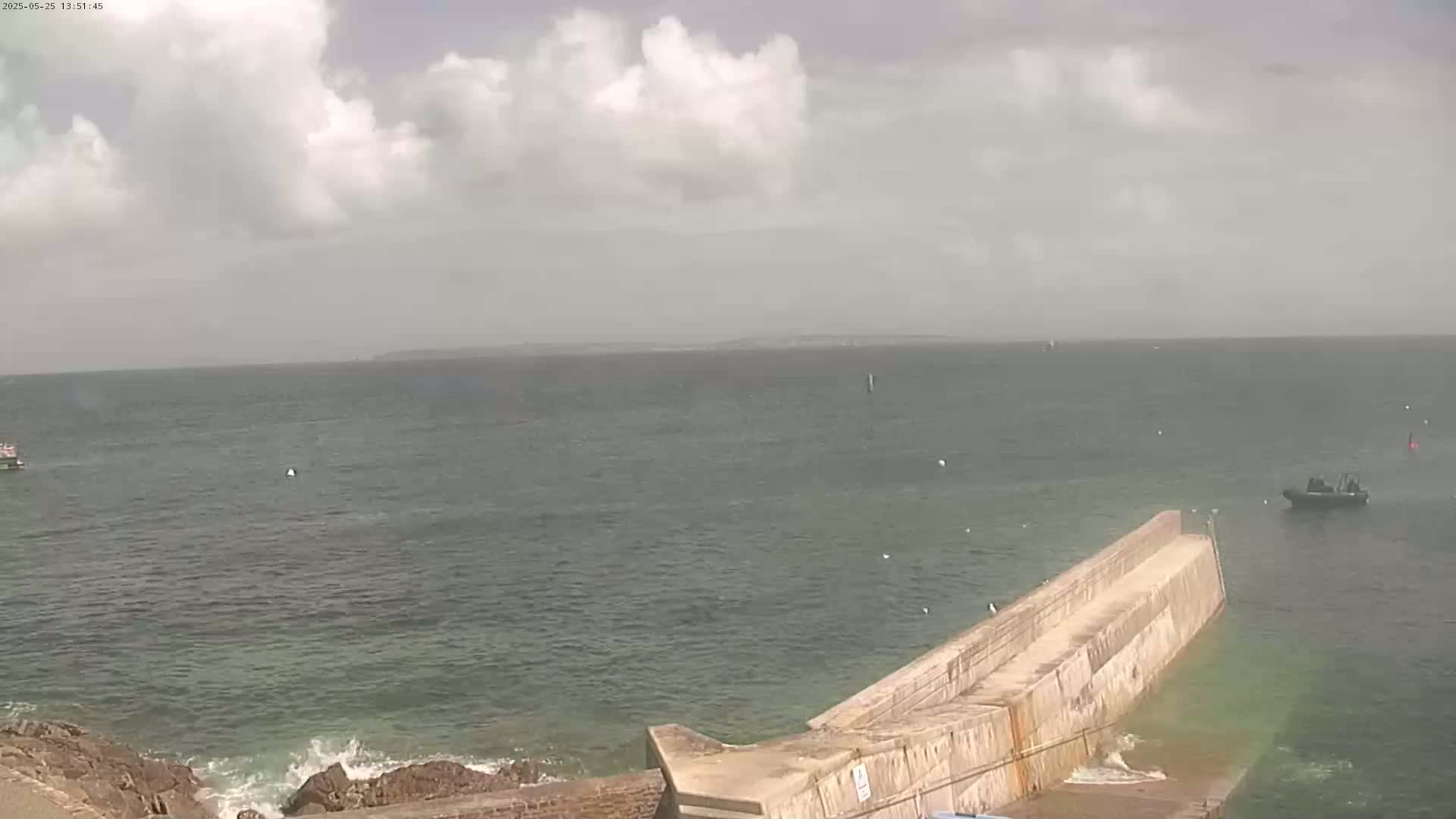 A partly cloudy day shows a wide expanse of calm sea with a few small boats, a long concrete jetty extending into the water from a rocky shoreline.