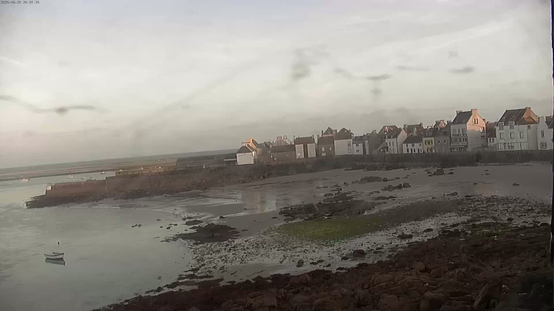 A row of buildings sits along a rocky shoreline next to calm water under an overcast sky.
