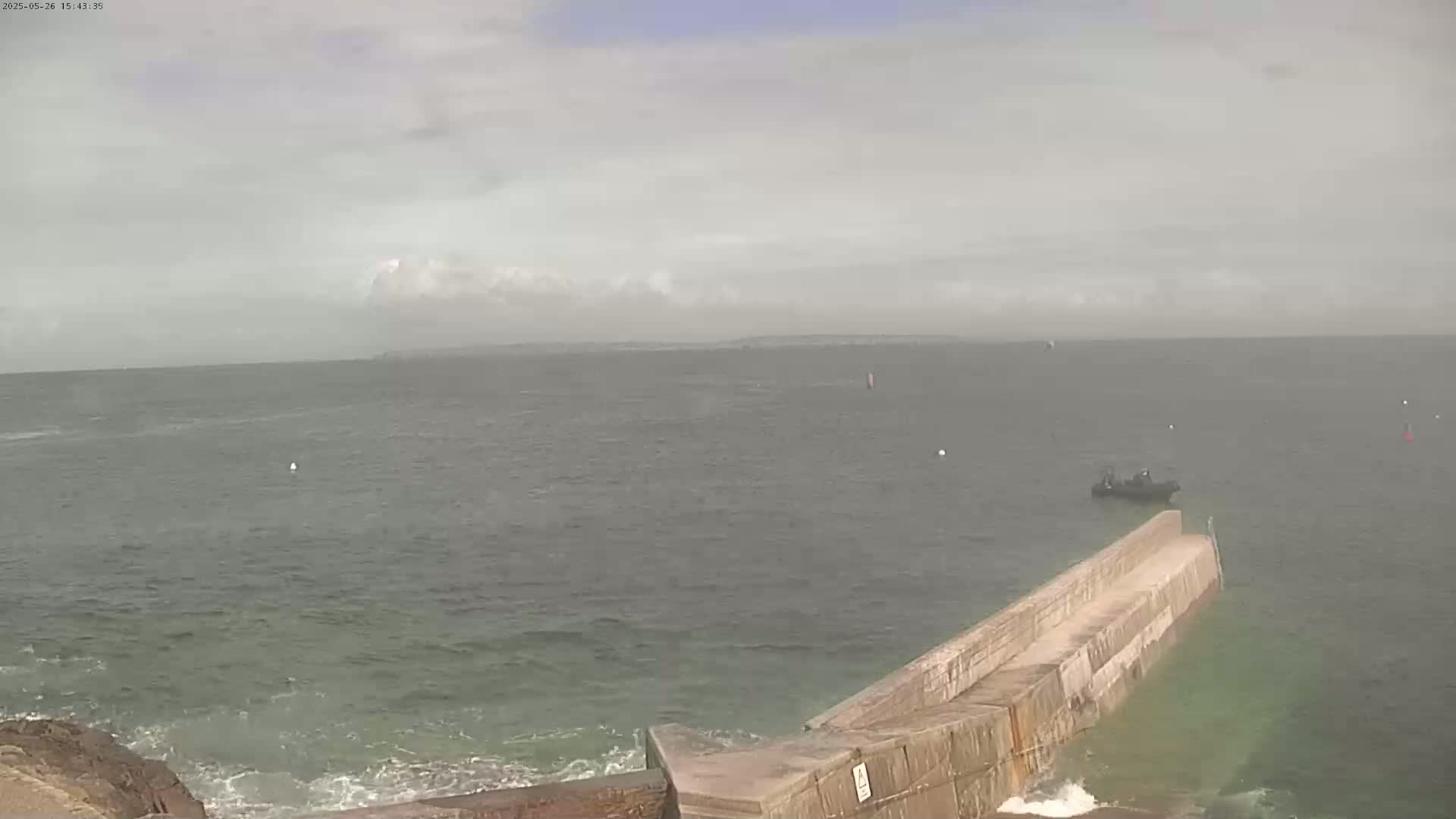 A partly cloudy day shows a body of water with a small boat near a long, low concrete structure extending into the ocean, with a hint of wave action near the shore.