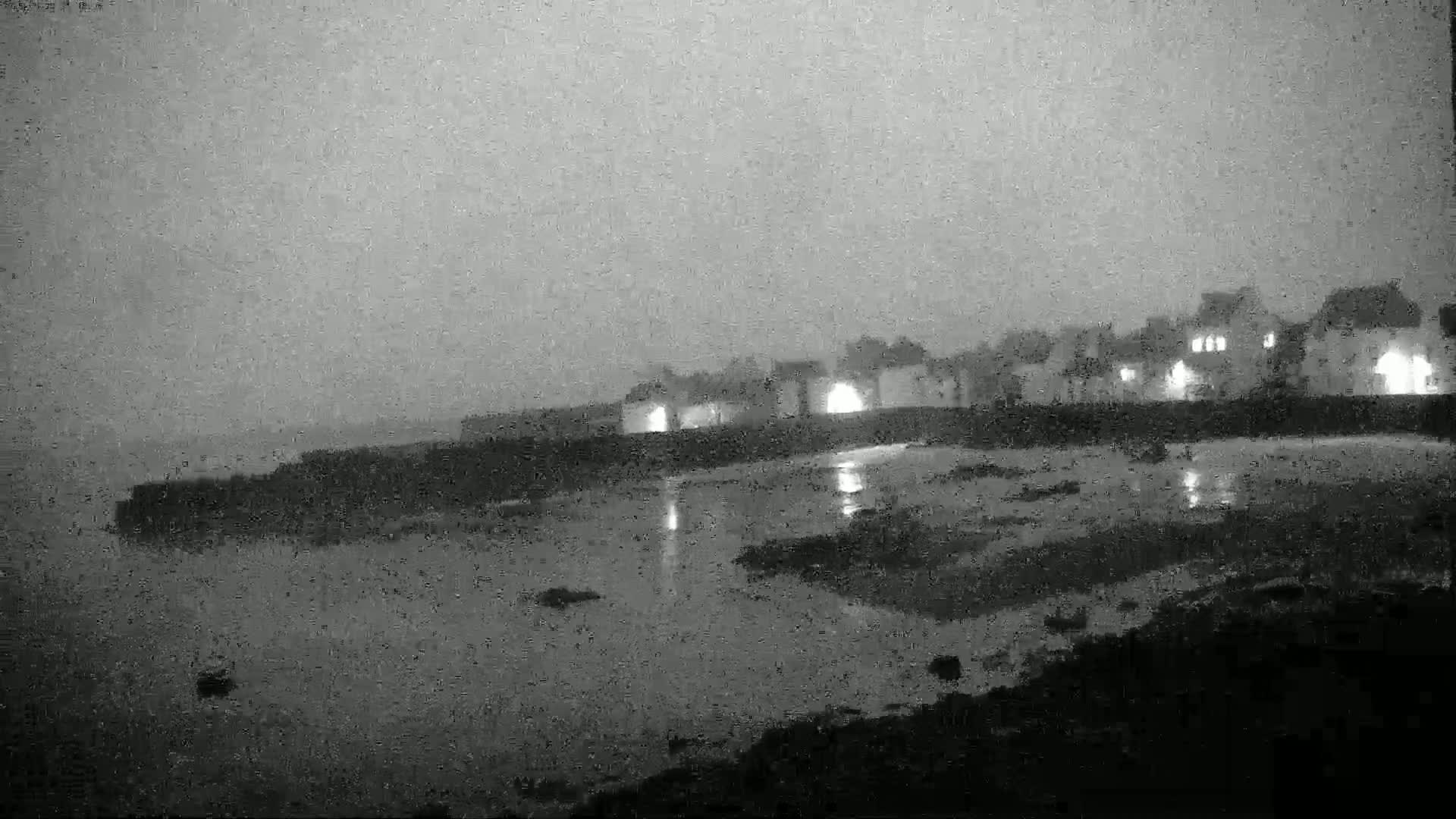 A dimly lit seaside town is seen at night under an overcast sky, with calm water reflecting the lights of the buildings.