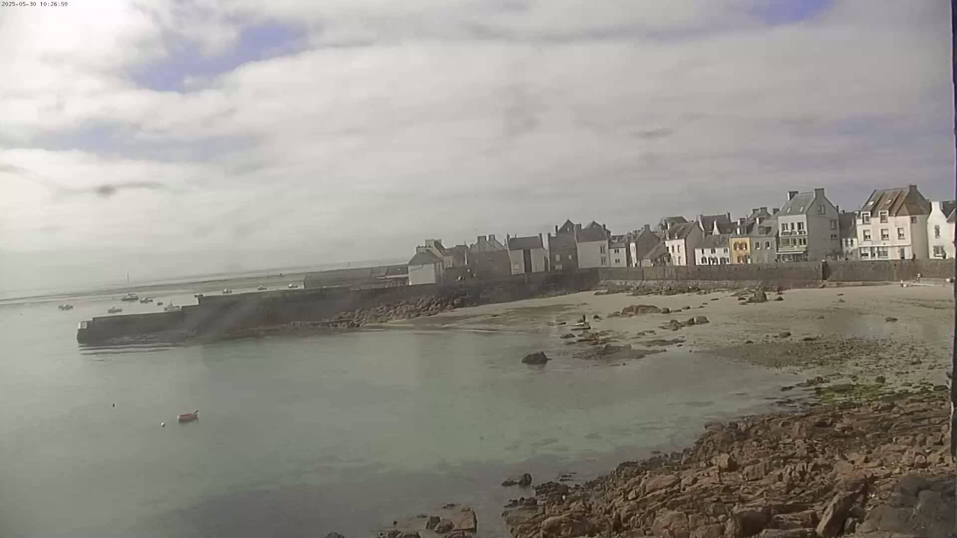 A row of buildings sits beside a sandy beach and calm, light-green water under a mostly cloudy sky.