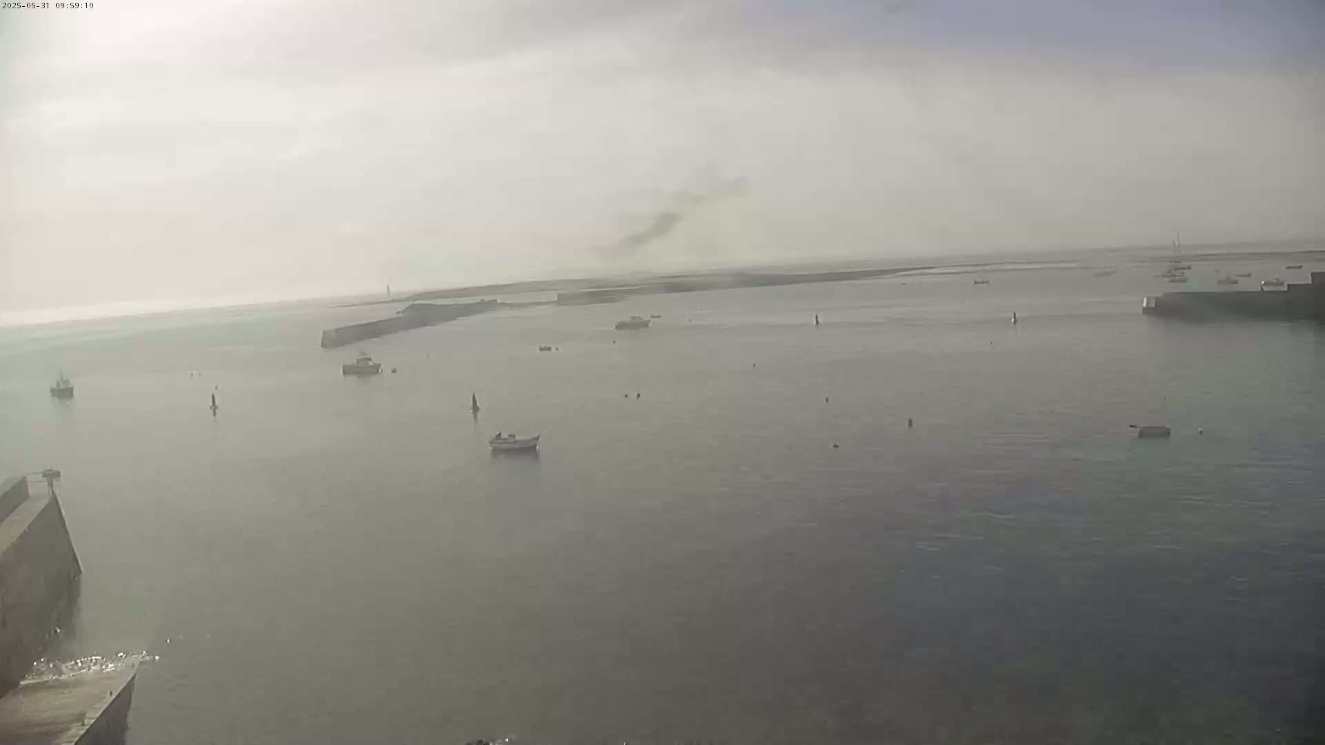 A hazy, overcast day reveals a harbor with several small boats, a stone jetty, and a distant shoreline.