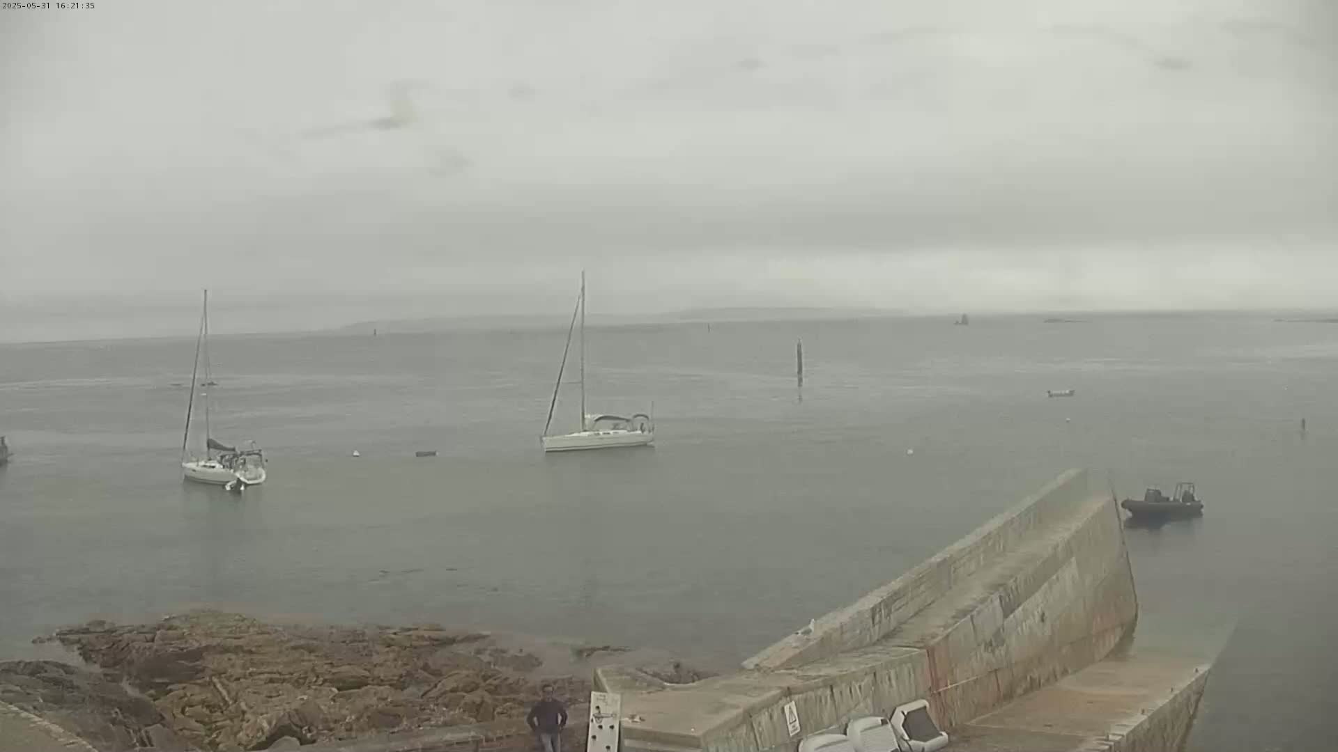 Several sailboats are anchored in a calm, foggy body of water near a rocky shore and a concrete ramp.
