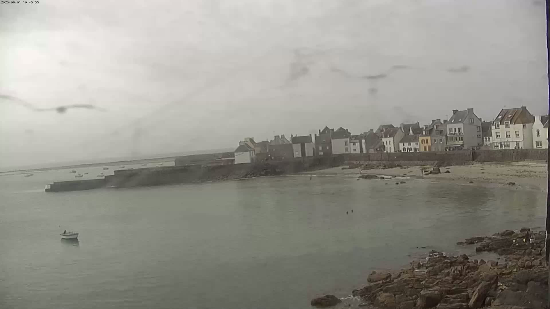 A small coastal town with a calm bay and a few boats is visible under an overcast, rainy sky.