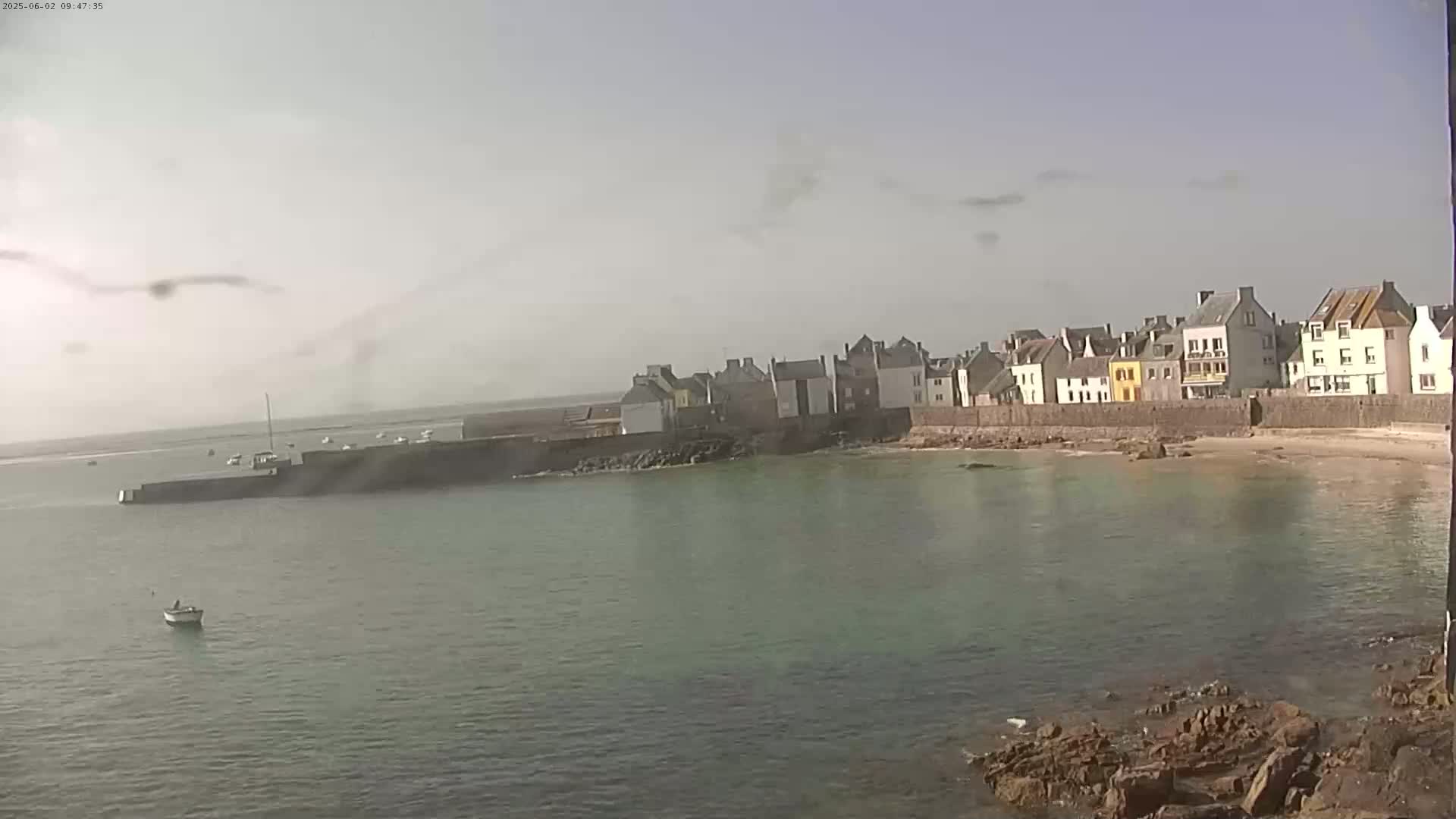 A small coastal town with multi-colored buildings sits on a rocky shoreline next to calm, light-green water under a hazy sky.
