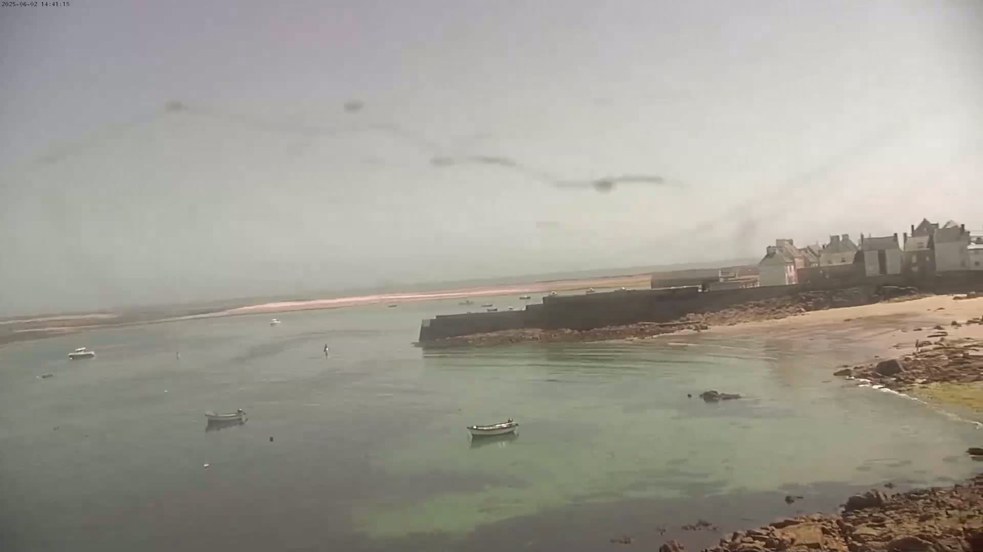 A hazy day reveals a calm bay with several small boats, a sandy beach, and a stone-walled village in the distance.