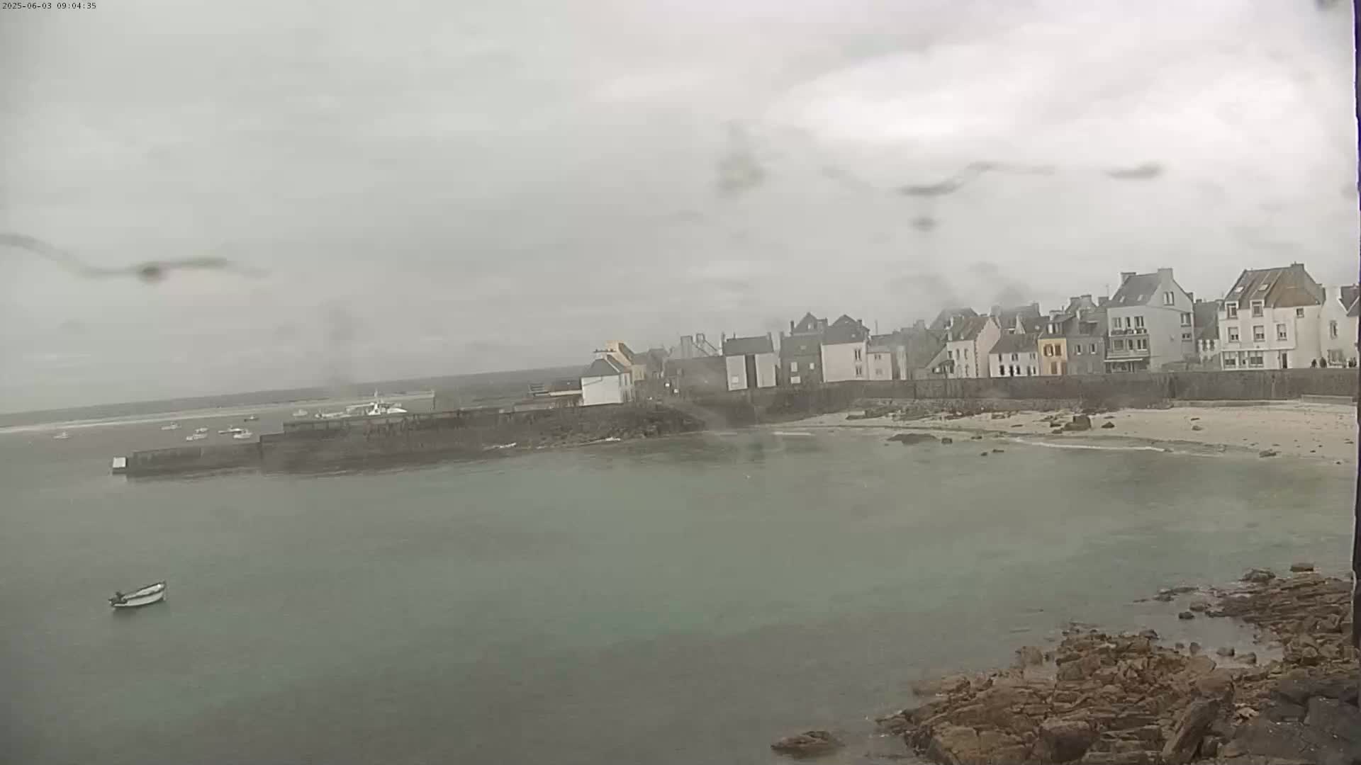 A small coastal town is seen on an overcast day, with a calm body of water in the foreground and several boats visible in the distance.