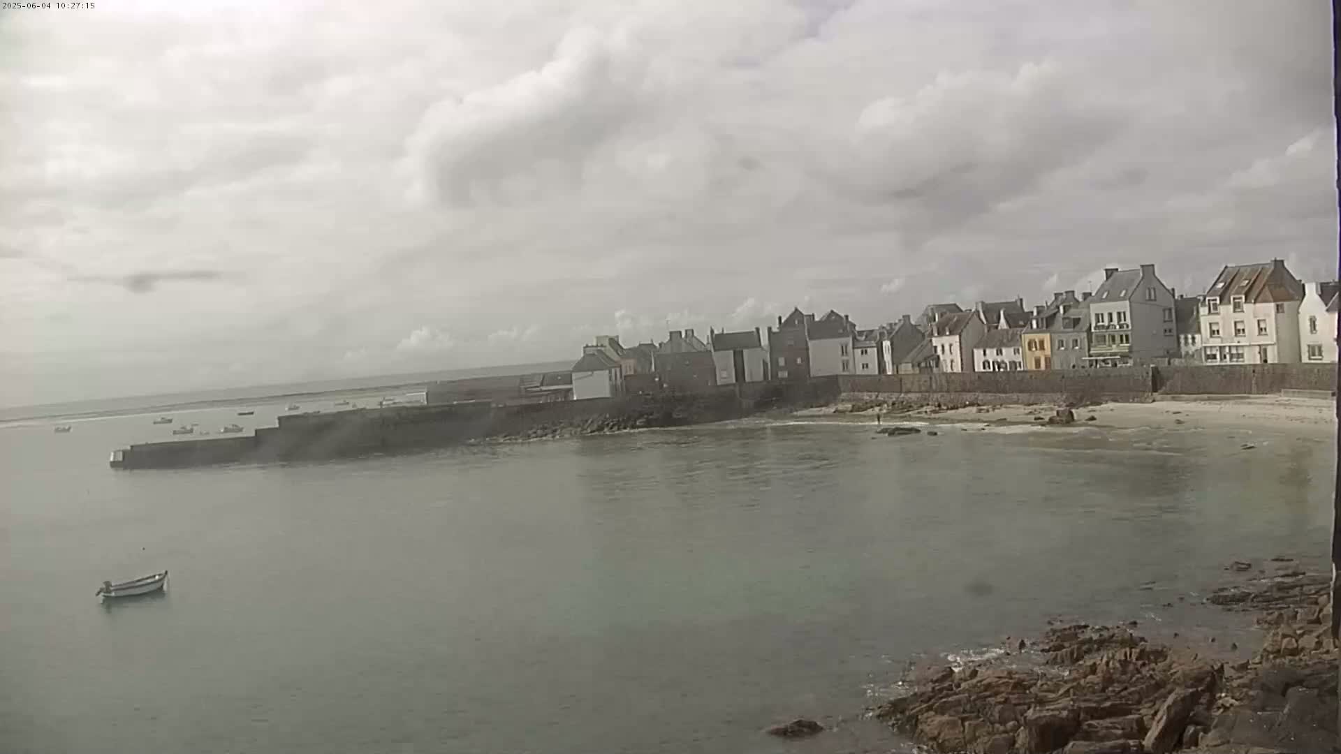 A small coastal town with several buildings sits on a calm body of water under a cloudy sky.