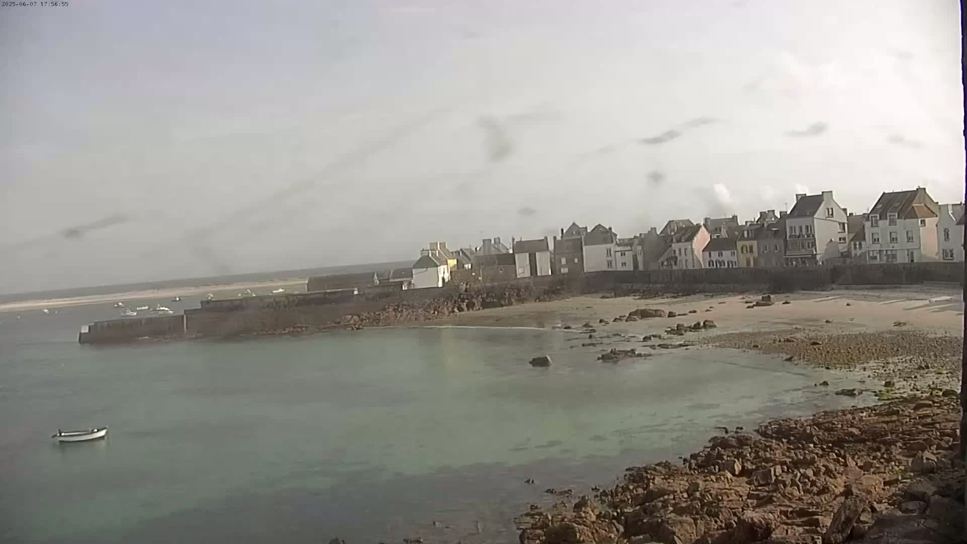 A small, coastal town with multi-story buildings sits beside a calm, light-green body of water under an overcast sky.