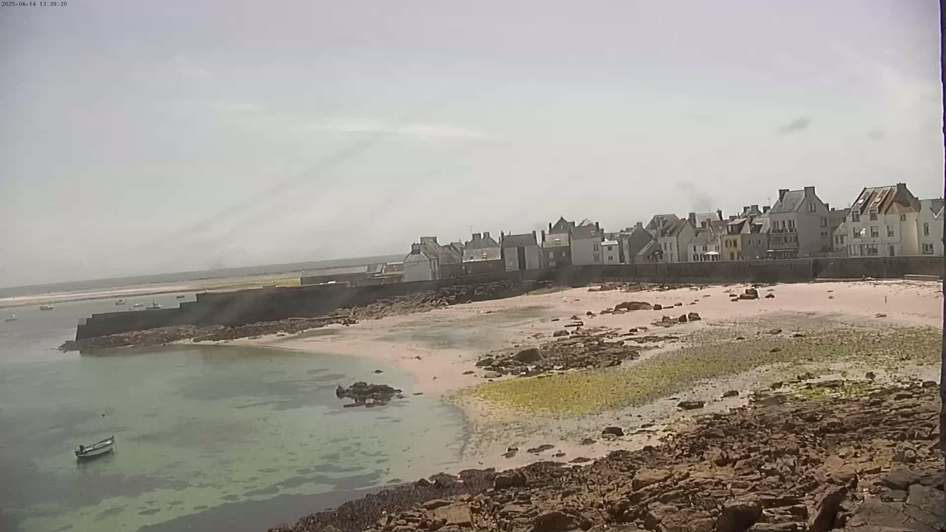 A coastal town with light-colored buildings sits behind a sandy beach and rocky shoreline on a hazy, sunny day.