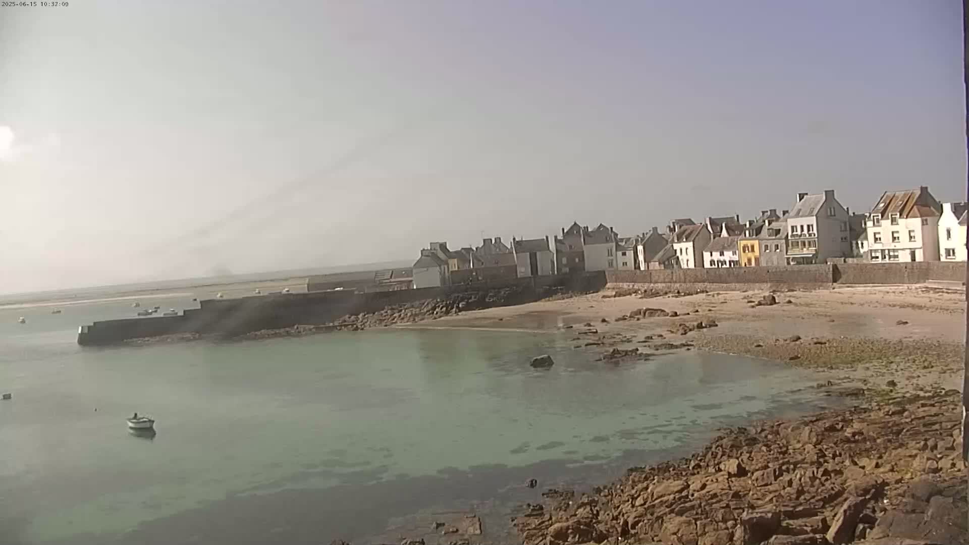 A hazy, sunny day reveals a coastal town with buildings along a low, sandy beach and a calm, shallow, green-tinged body of water.