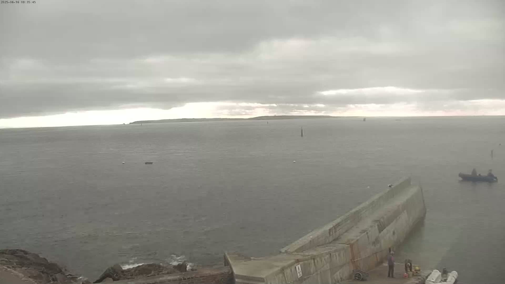 A gray, overcast sky hangs over a calm body of water, where a small boat is visible near a long concrete pier extending from a rocky shoreline.
