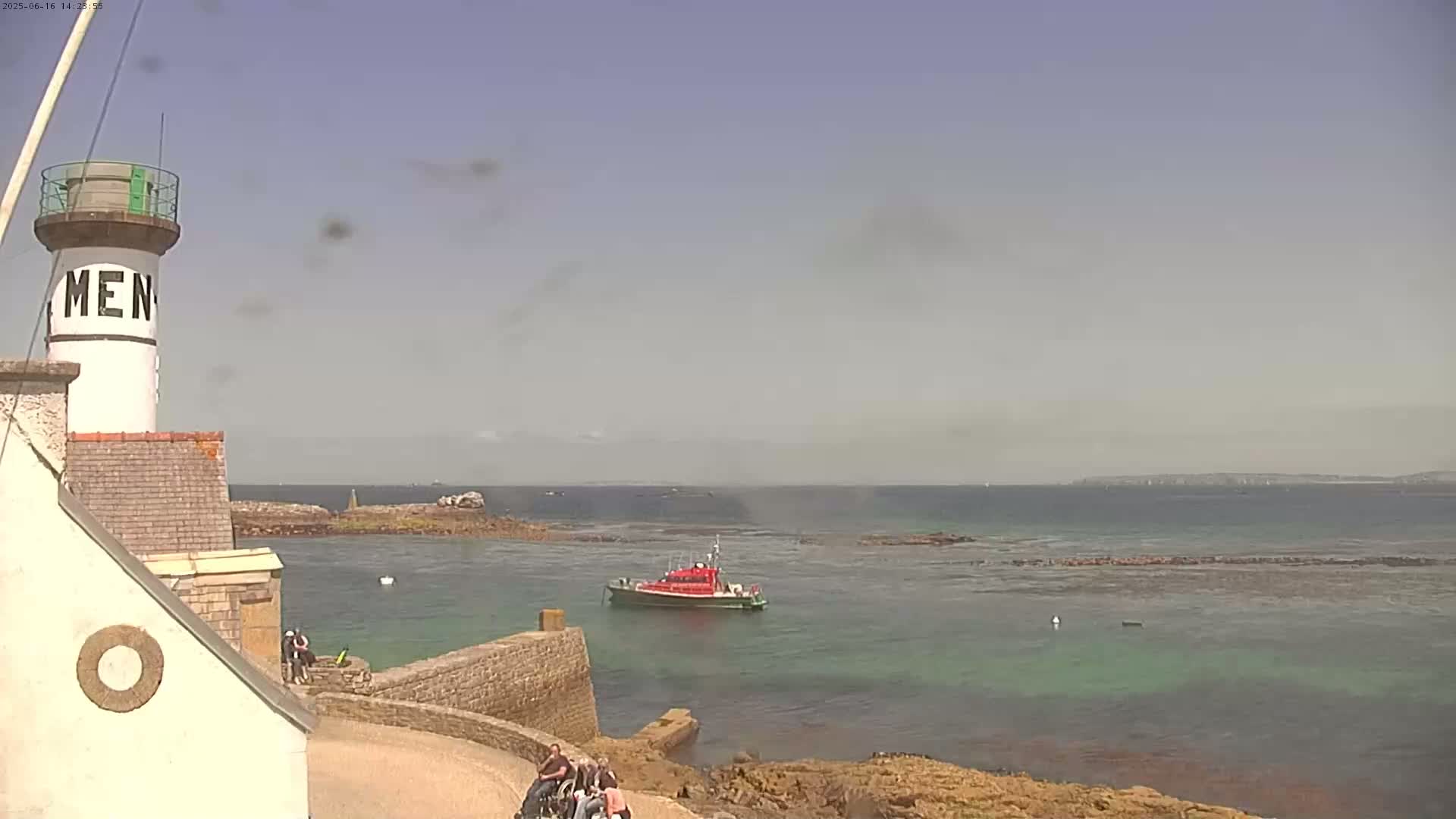 A white lighthouse with a red lifeboat in the calm, hazy waters near a rocky shoreline.
