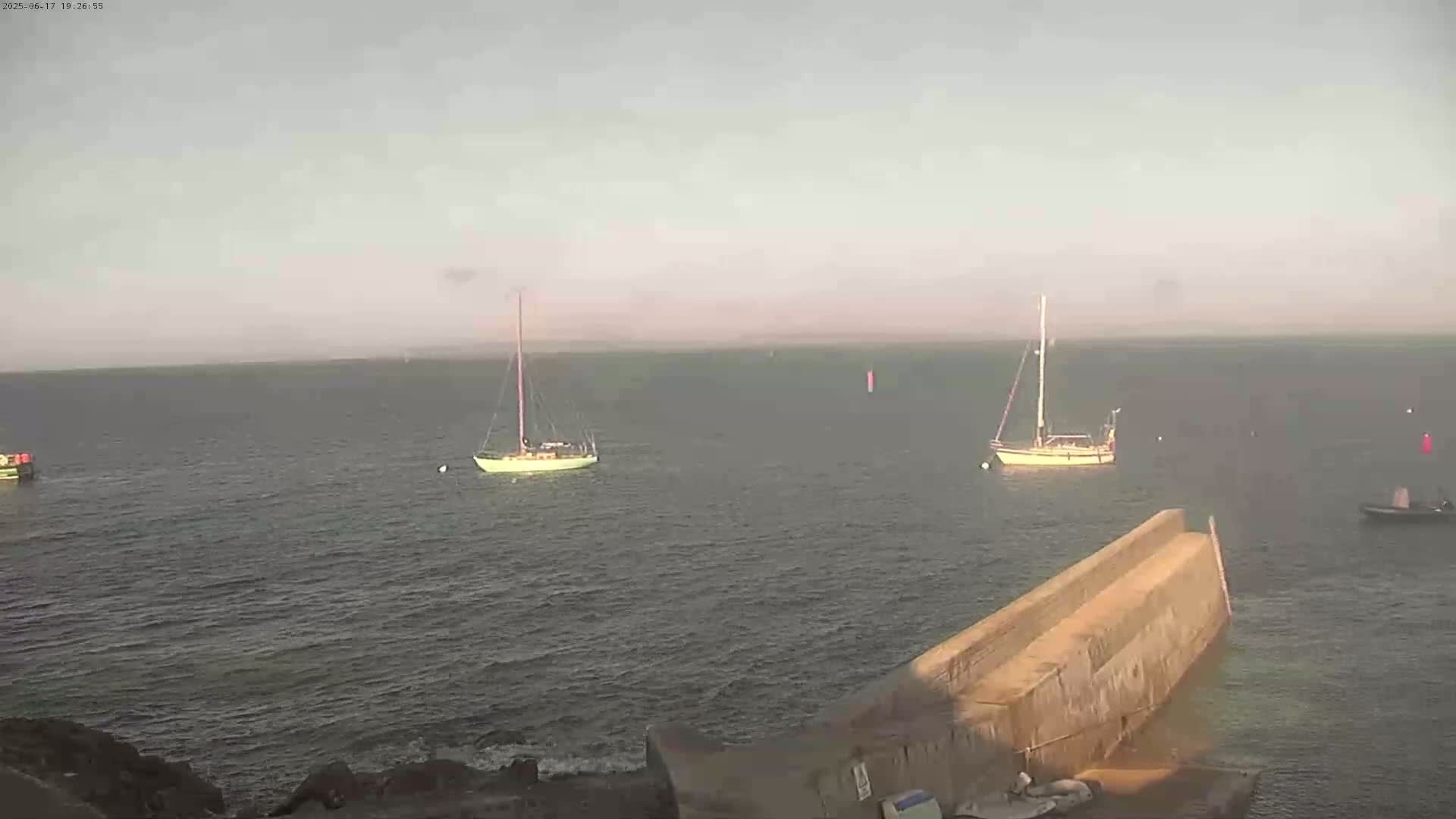 Several sailboats are anchored in calm water near a stone breakwater under an overcast sky.
