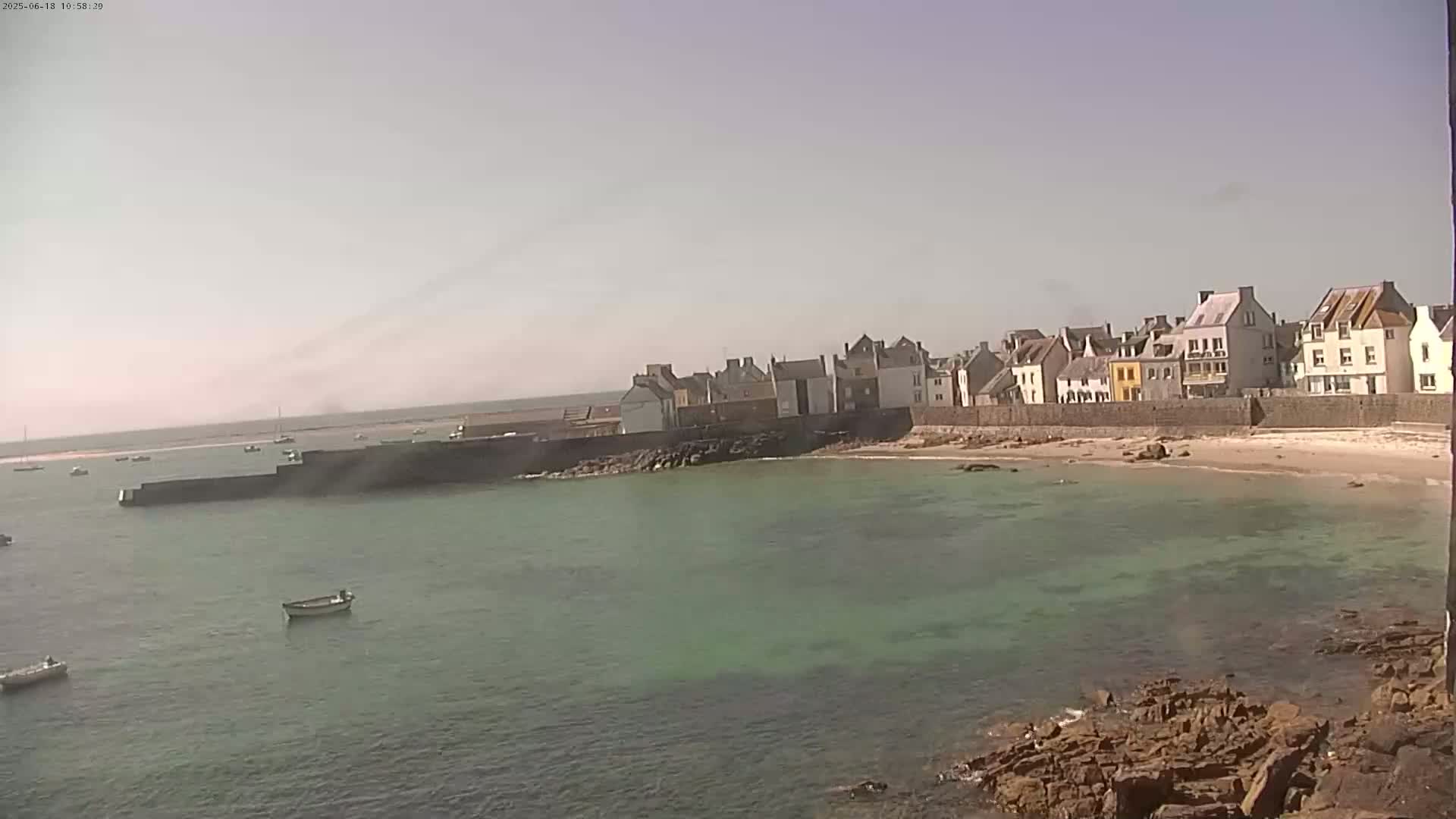 A quaint coastal village with a small sandy beach and calm, shallow green water sits under a hazy sky.