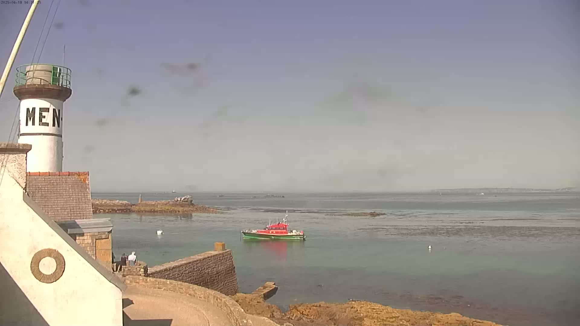 A lighthouse stands on a rocky coast next to calm, hazy water where a small boat is anchored; a few people are visible near the lighthouse.