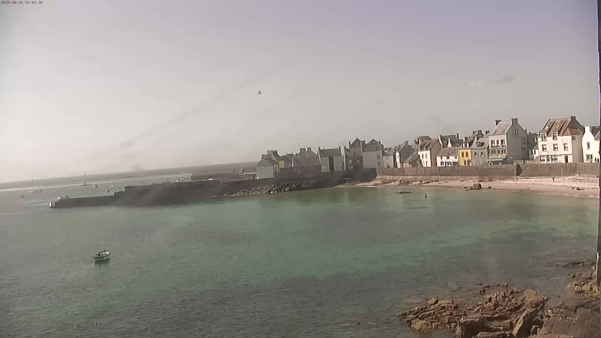 A small coastal town with light-colored buildings is situated along a calm, greenish body of water on a hazy day.