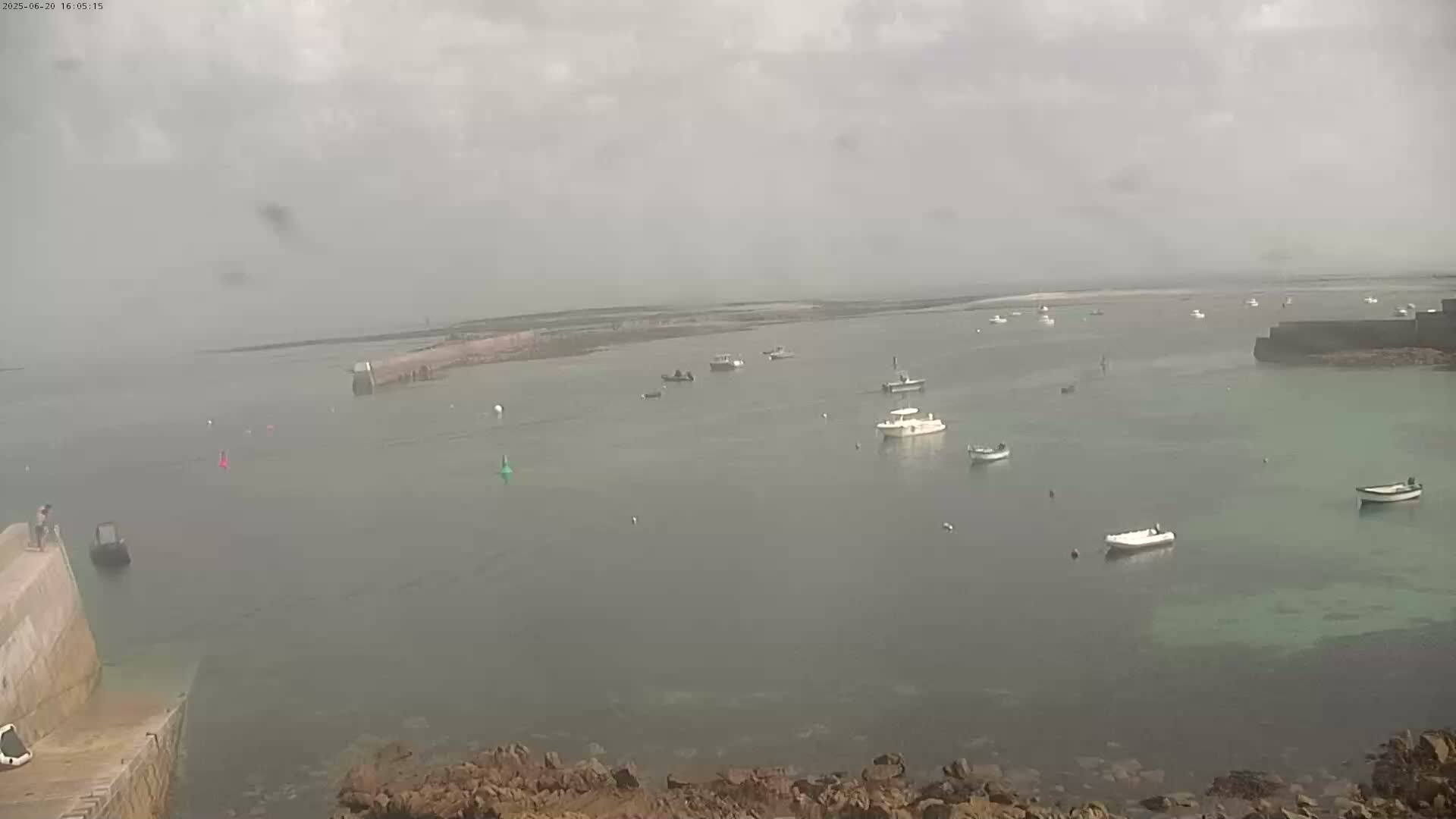 A hazy, overcast day reveals a calm harbor with numerous small boats anchored in shallow, greenish water, partially enclosed by rocky land.