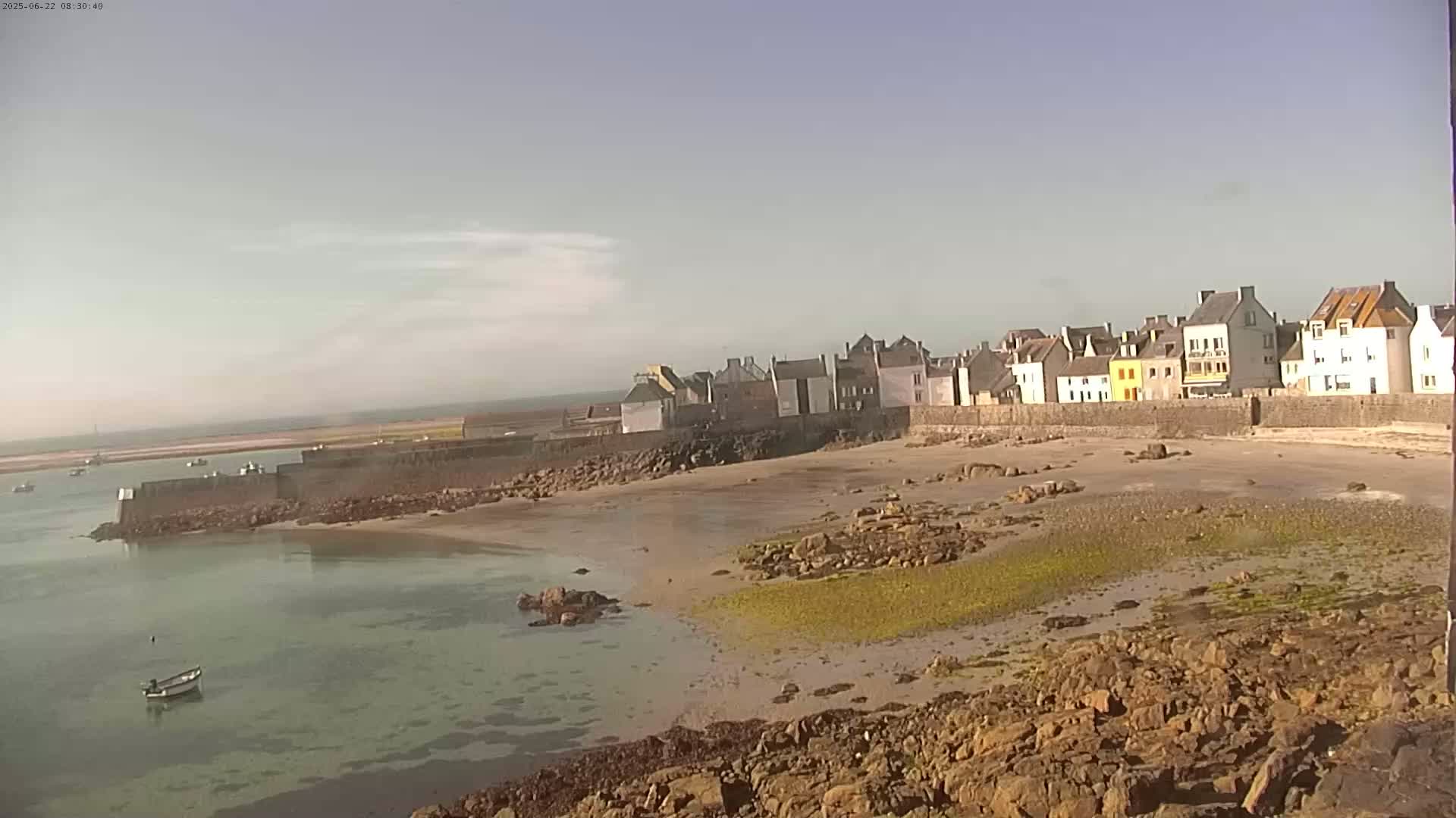 A small coastal town with stone buildings sits behind a sandy beach and rocky shoreline on a partly sunny day.