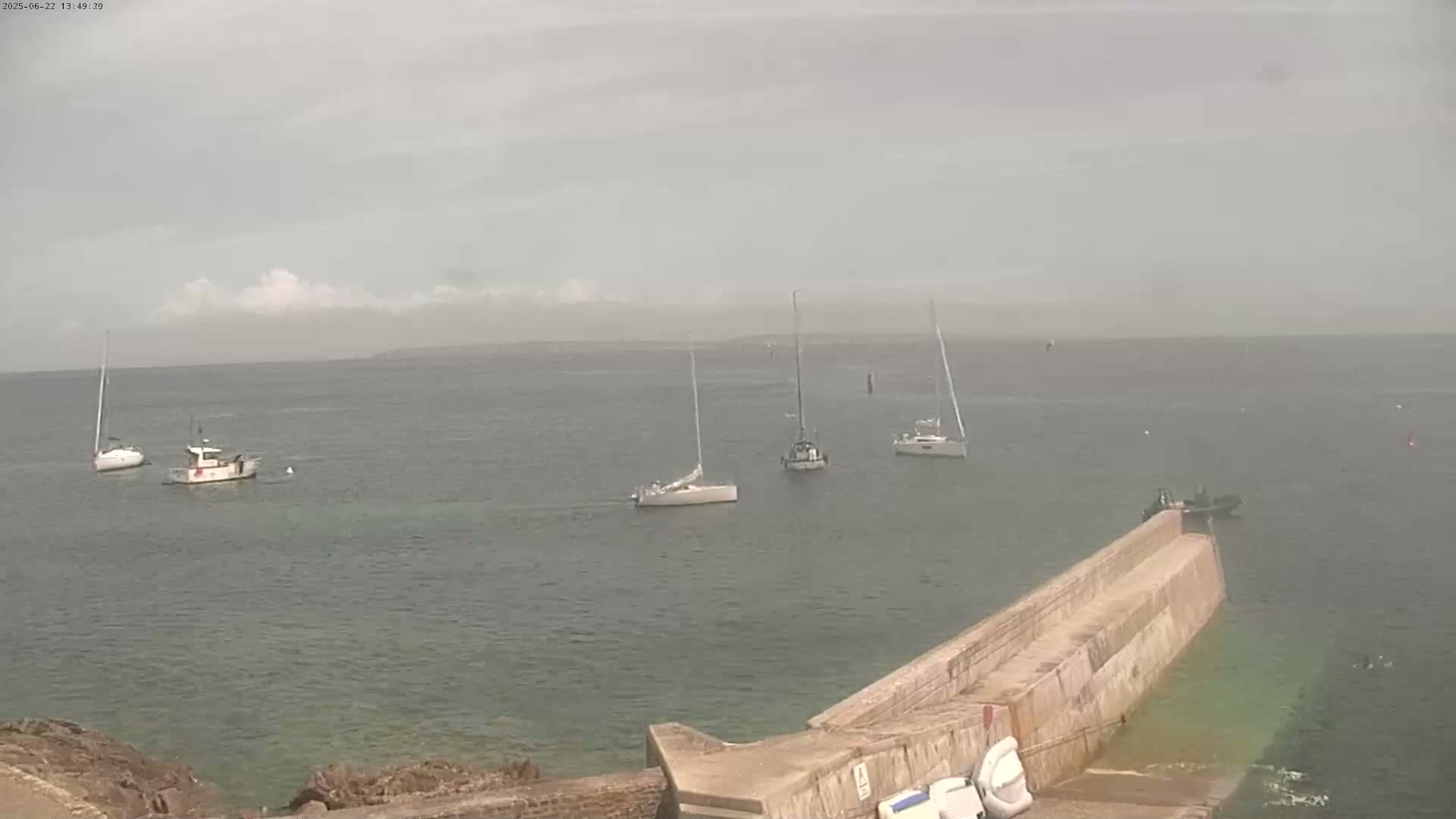 Several sailboats and motorboats are anchored in calm, overcast waters near a concrete pier.