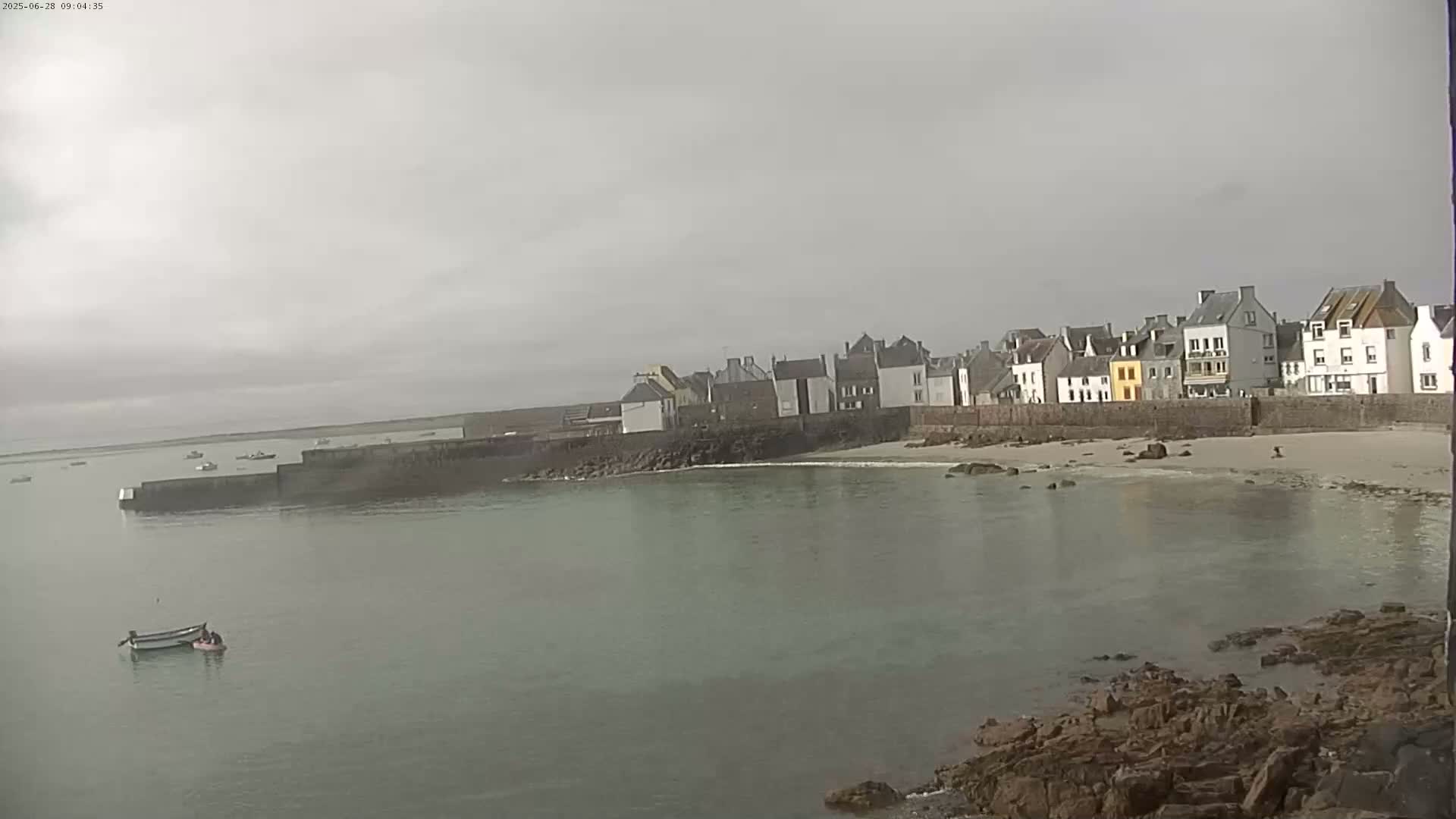 A small coastal town with several buildings is seen across a calm, greenish-gray body of water under an overcast sky.