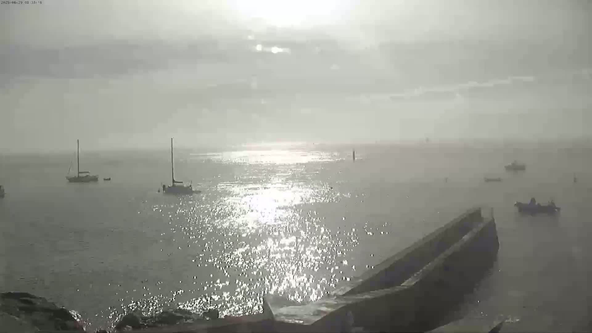 A hazy, sunlit ocean scene shows several sailboats and motorboats, with a low stone wall or jetty in the foreground.