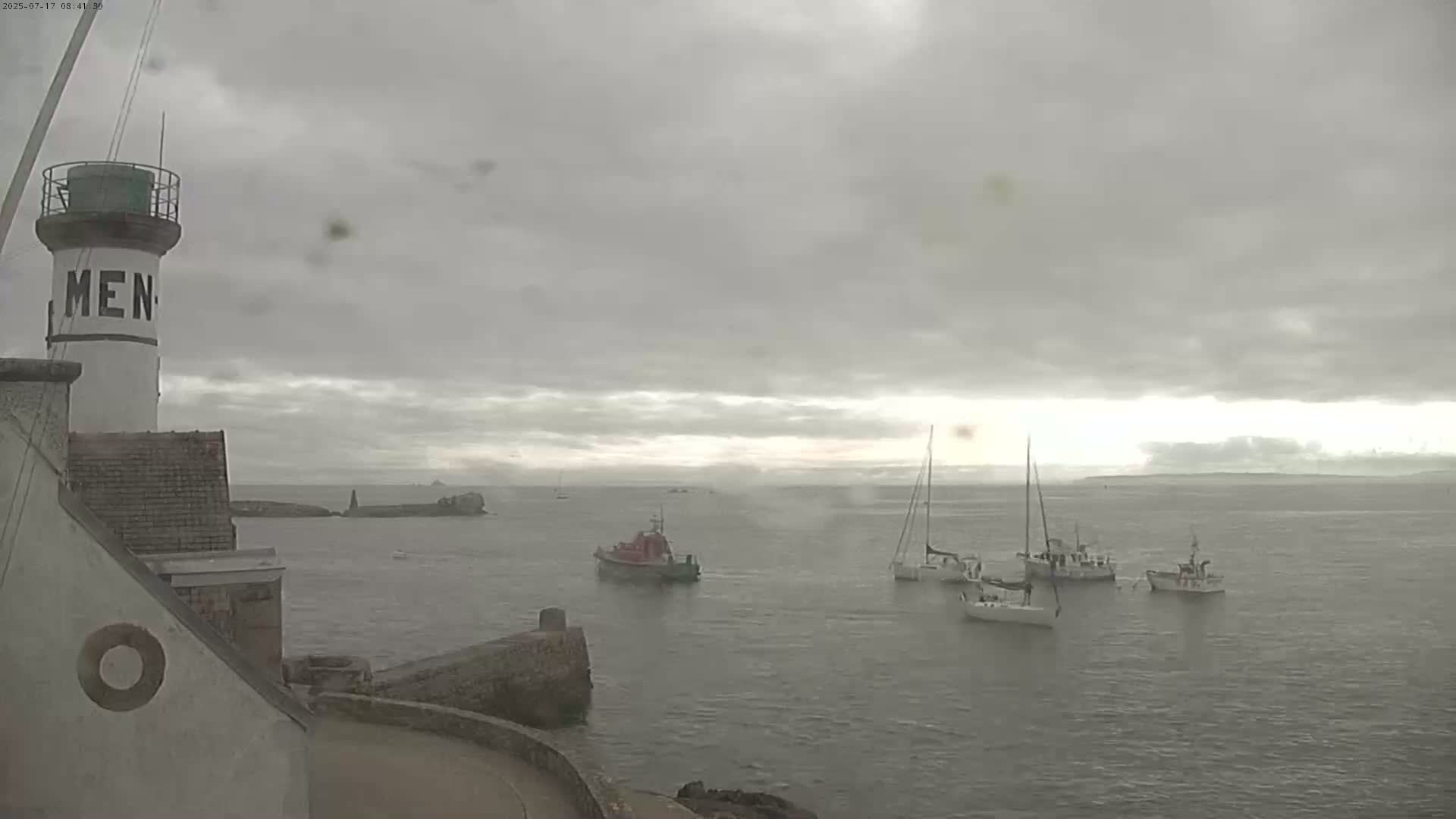 A lighthouse stands beside a calm body of water under an overcast sky, with several boats visible in the distance.