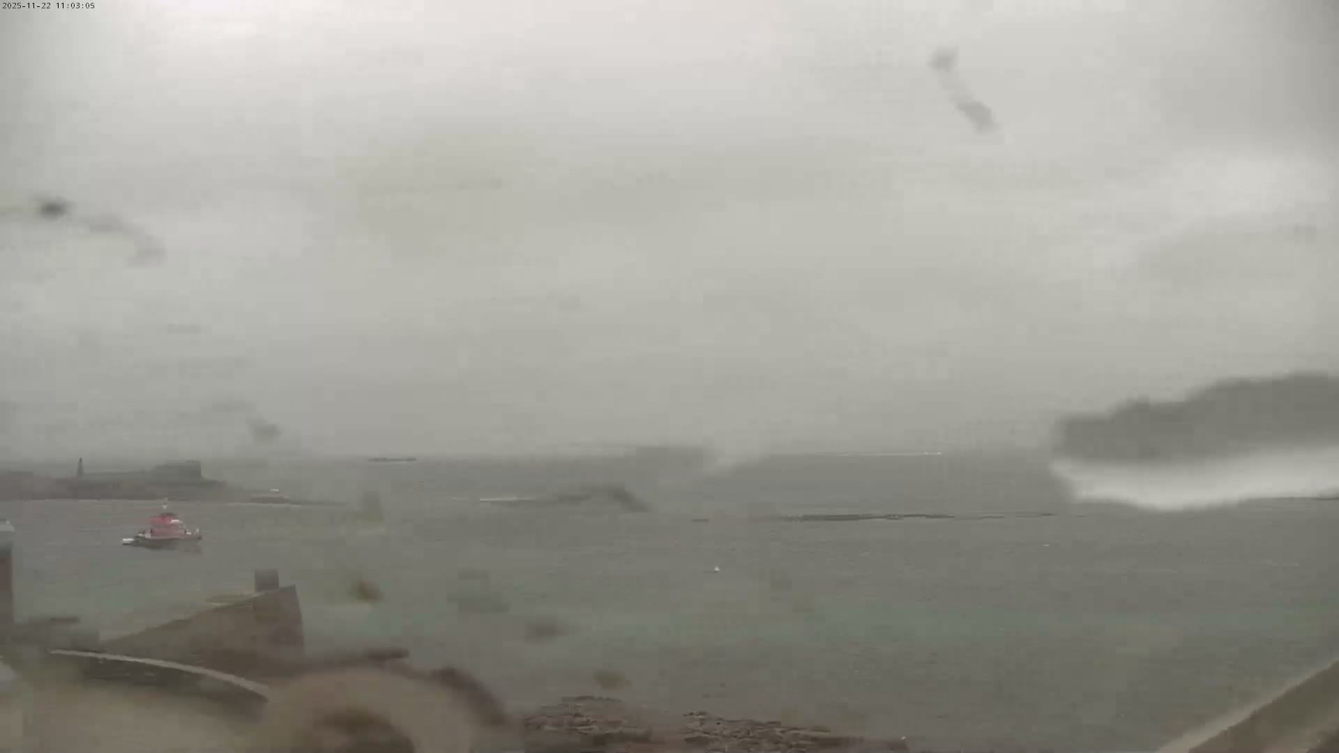 A red and white tugboat navigates choppy, dark gray waters under a heavily overcast and rainy sky, with distant land and structures appearing hazy in the low visibility.