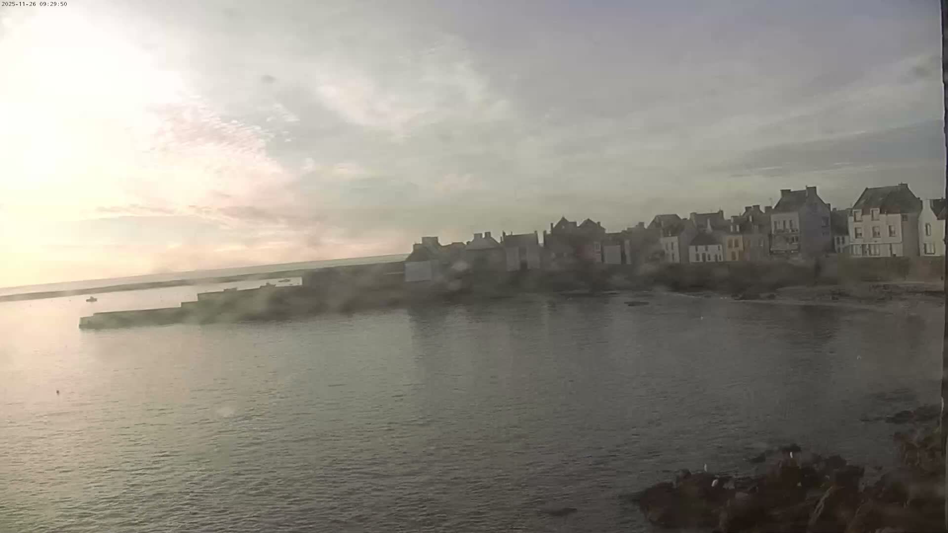Under a hazy and partly cloudy sky, a coastal town with traditional buildings lines the shore of a calm bay, featuring a pier and a distant boat.