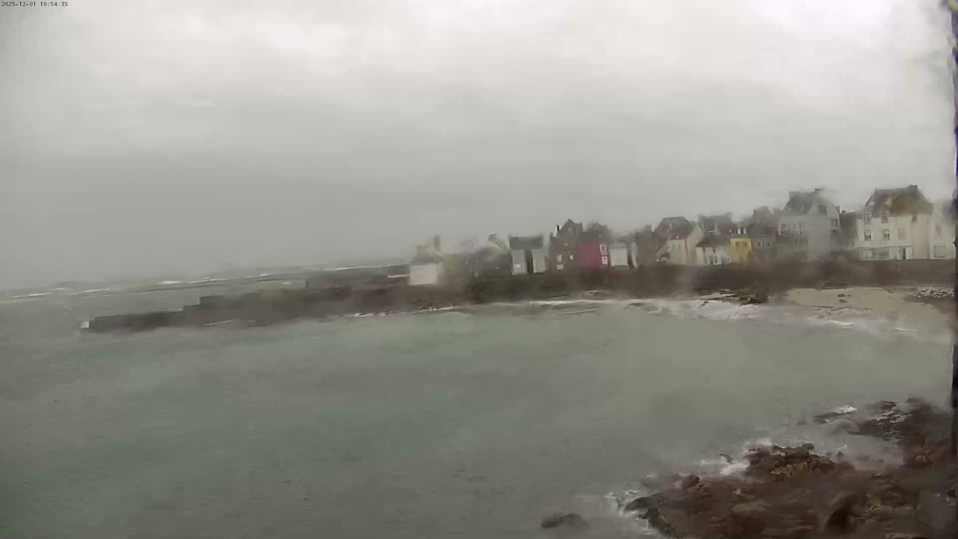 A wide, blurry view captures a line of buildings along a rocky coastline with choppy grey-green water and breaking waves under a heavily overcast, stormy sky, suggesting rain or mist.
