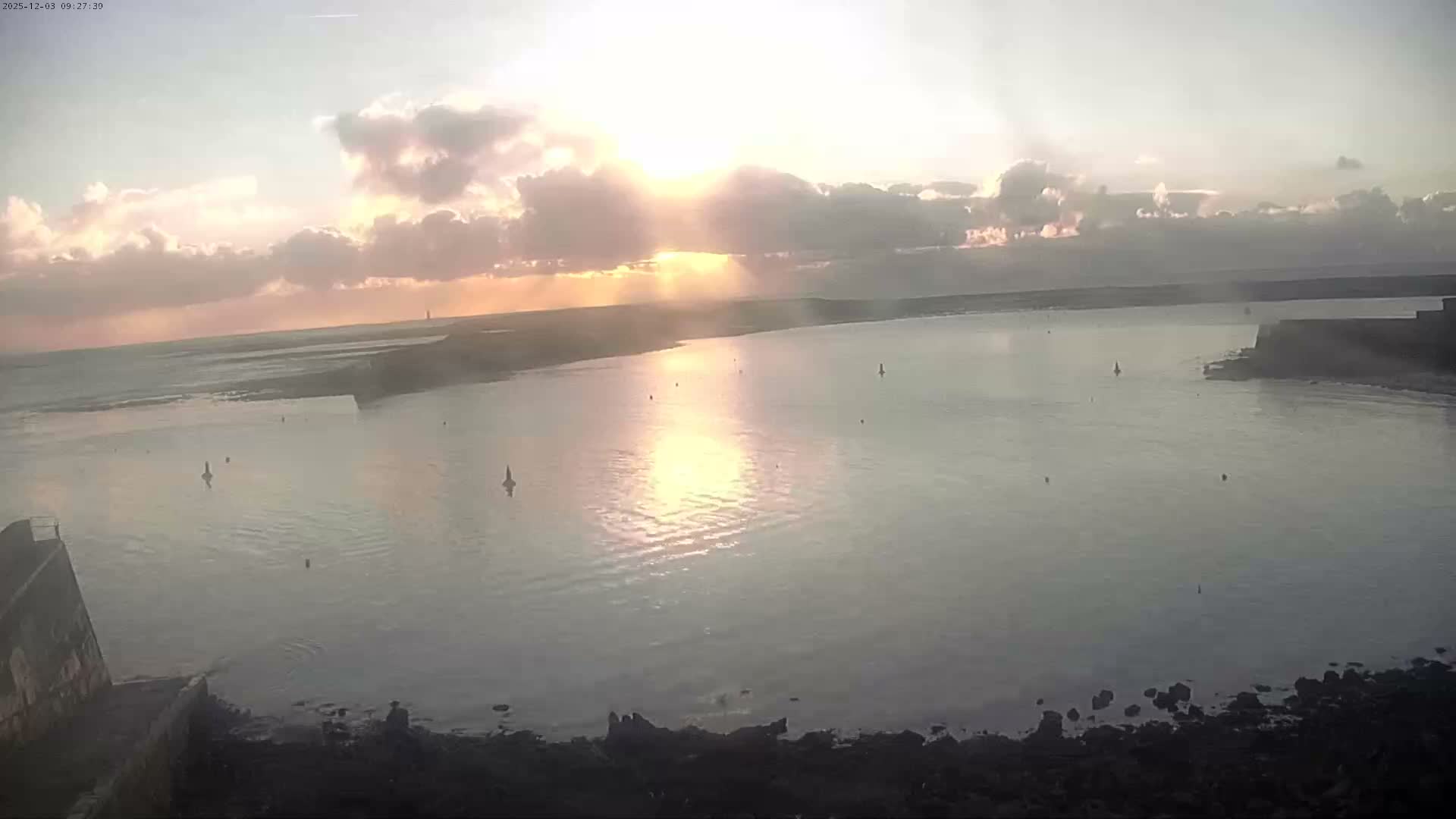 A tranquil bay or estuary is illuminated by brilliant, low-lying sun rays breaking through scattered clouds, casting a golden reflection on the calm water where several small buoys are visible near the shorelines.