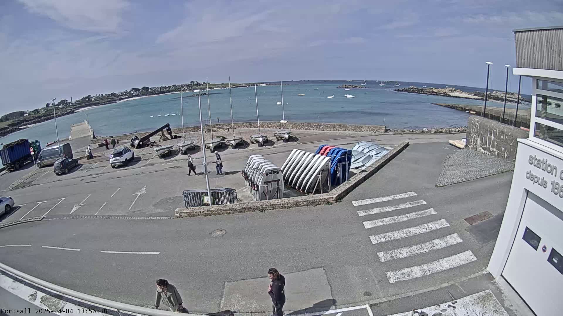 A sunny day at a harbor shows several parked cars, sailboats, and people walking near a building and calm, light blue water.