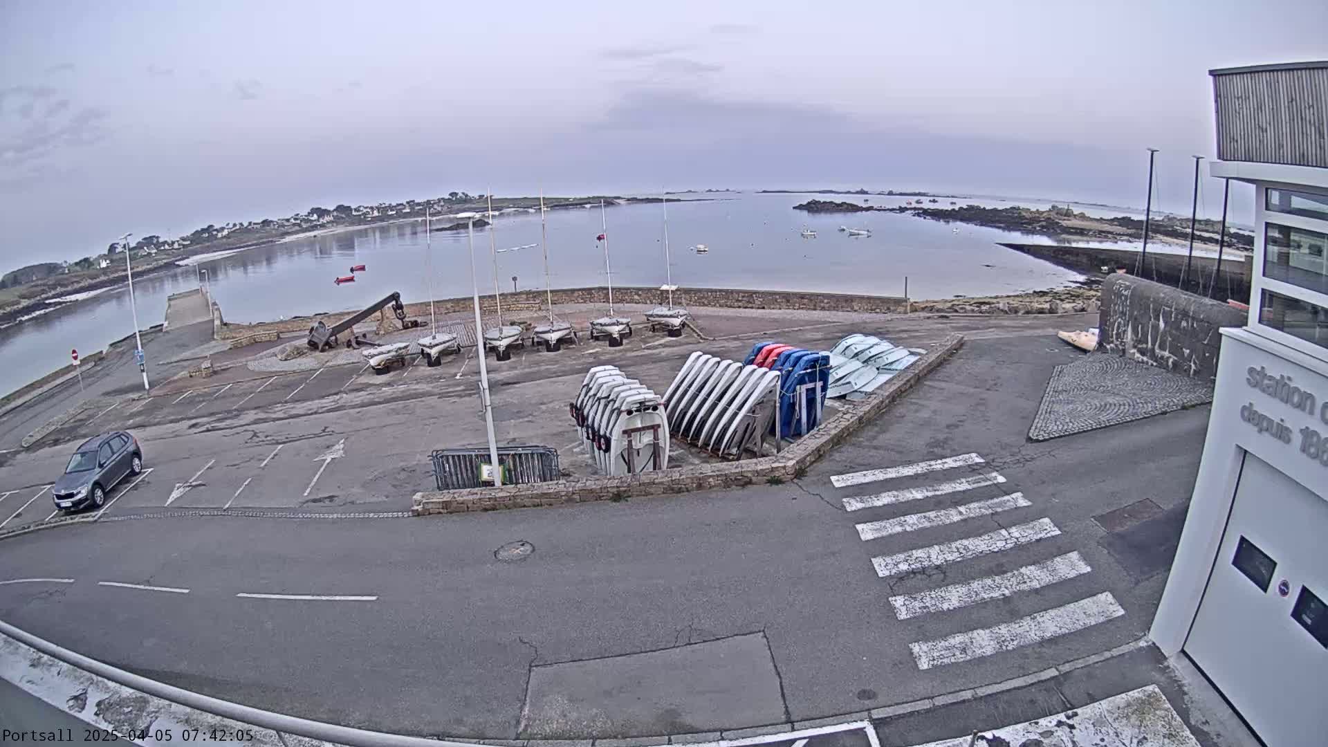 An overcast day shows a harbor with several sailboats and dinghies docked, a car in a parking lot, and a building.