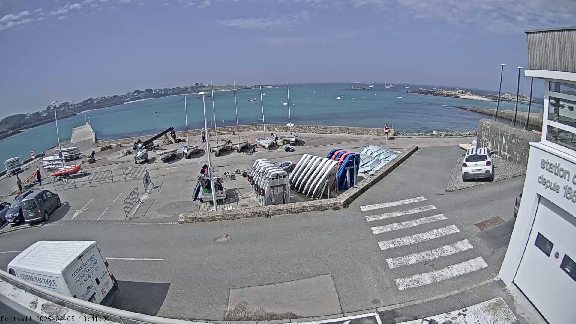 A sunny day at a harbor with numerous sailboats and small motorboats docked, along with several cars parked in a lot overlooking a calm body of water.
