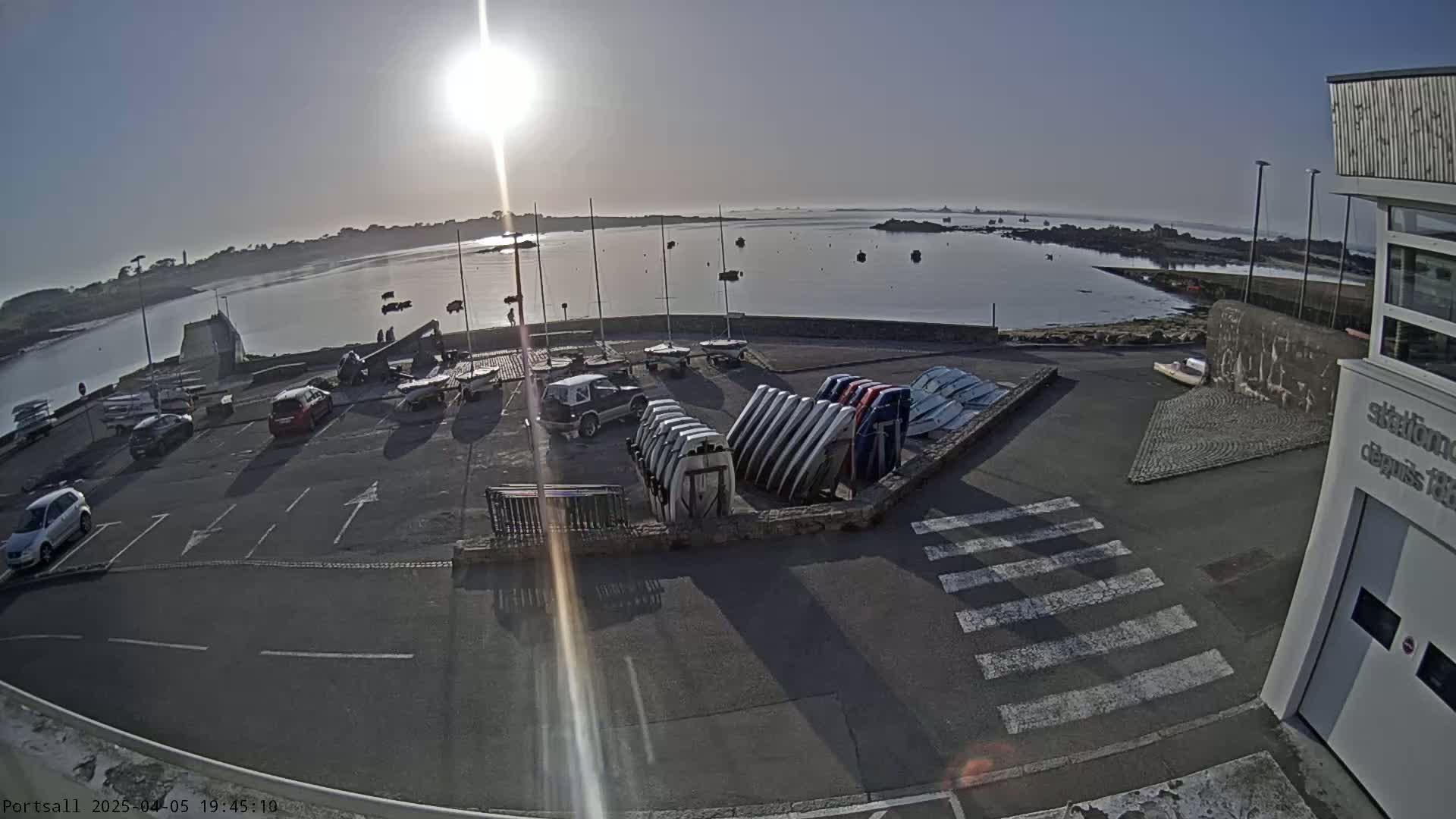 A sunny morning view of a harbor with several parked cars and boats, along with a building and a crosswalk in the foreground.