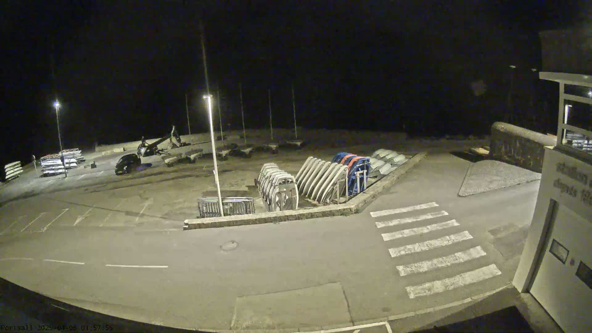 A nighttime, outdoor view of a paved area with parked cars, stacked boats, and a pedestrian crossing.
