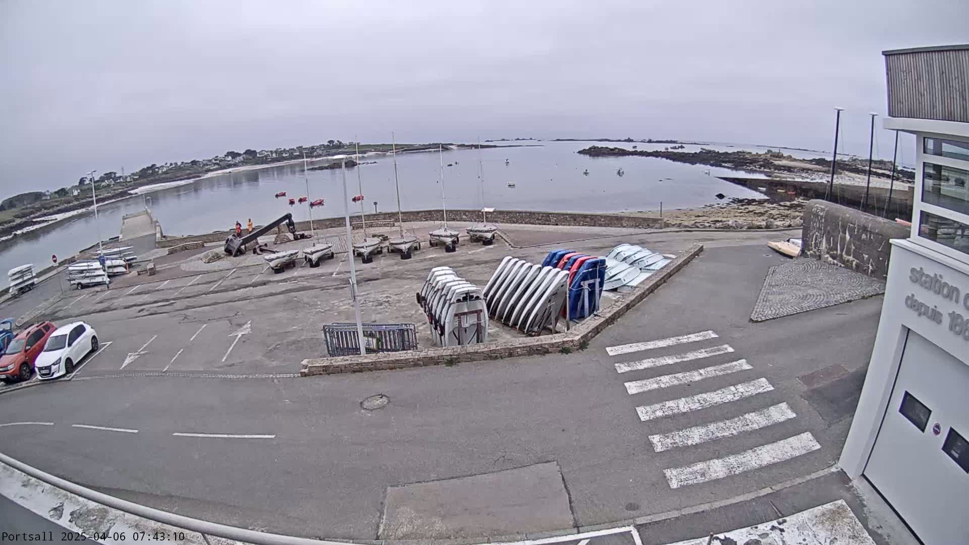 A harbor with numerous small boats and sailboats on land and in calm, overcast water, next to a parking lot and a building.